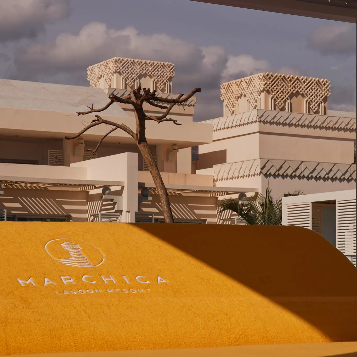 Un bâtiment beige avec des balcons et des motifs géométriques sur les toits, sous un ciel partiellement nuageux, avec un panneau "Marchica Lagoon Resort" en premier plan