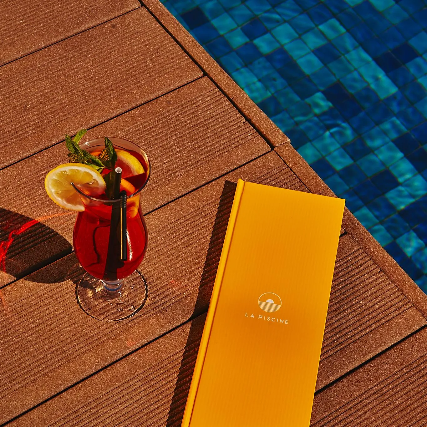 Un verre de boisson rouge avec une tranche d'orange et une paille, un menu jaune et une piscine bleue en arrière-plan