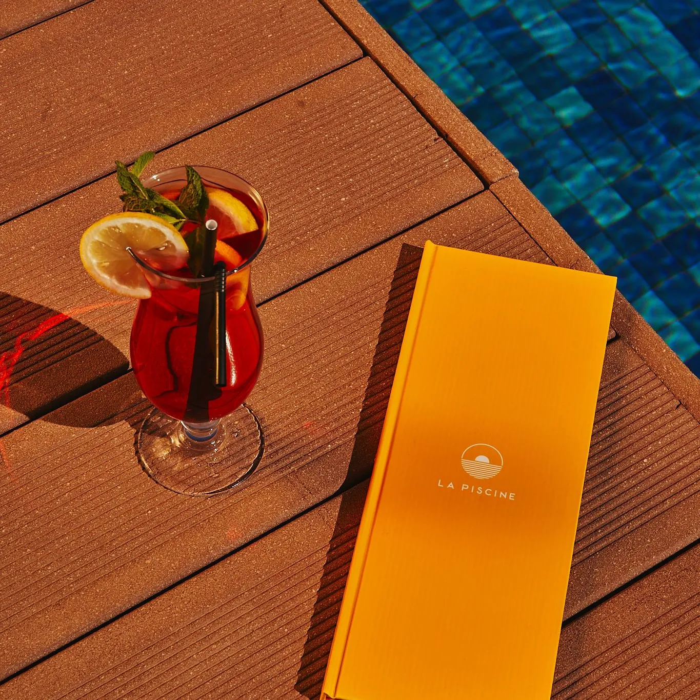 Un verre de boisson rouge avec une tranche d'orange et une paille, un menu orange et une piscine bleue en arrière-plan