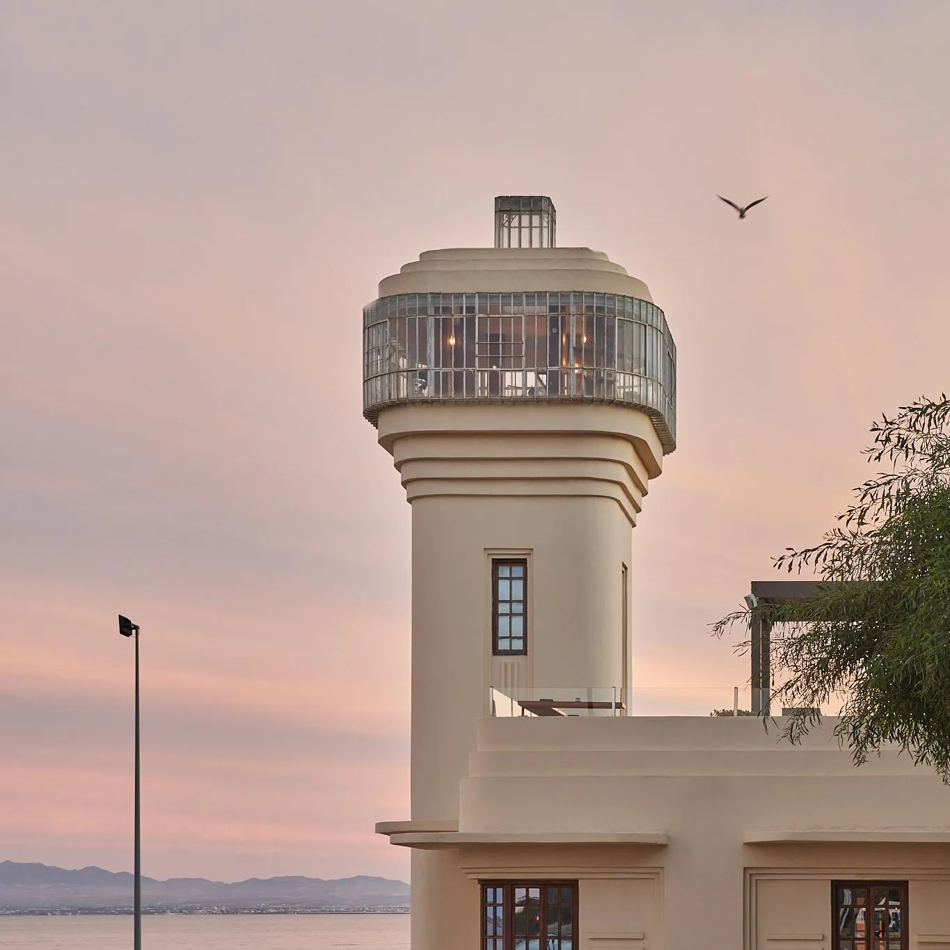 Un phare moderne avec une lanterne vitrée en haut se dresse près d'une étendue d'eau au coucher du soleil