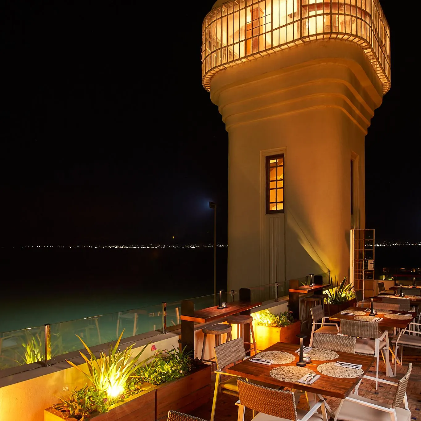 Terrasse de restaurant éclairée la nuit avec tables, chaises et plantes, située près d'une structure cylindrique illuminée et d'une piscine