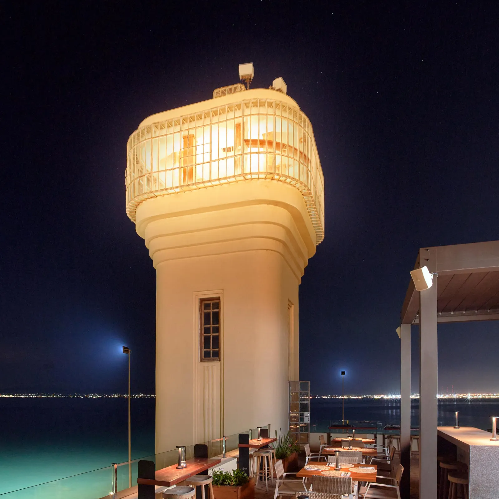 Une terrasse de restaurant en bord de mer avec des tables et des chaises, éclairée la nuit, située près d'une tour avec une structure en treillis illuminée