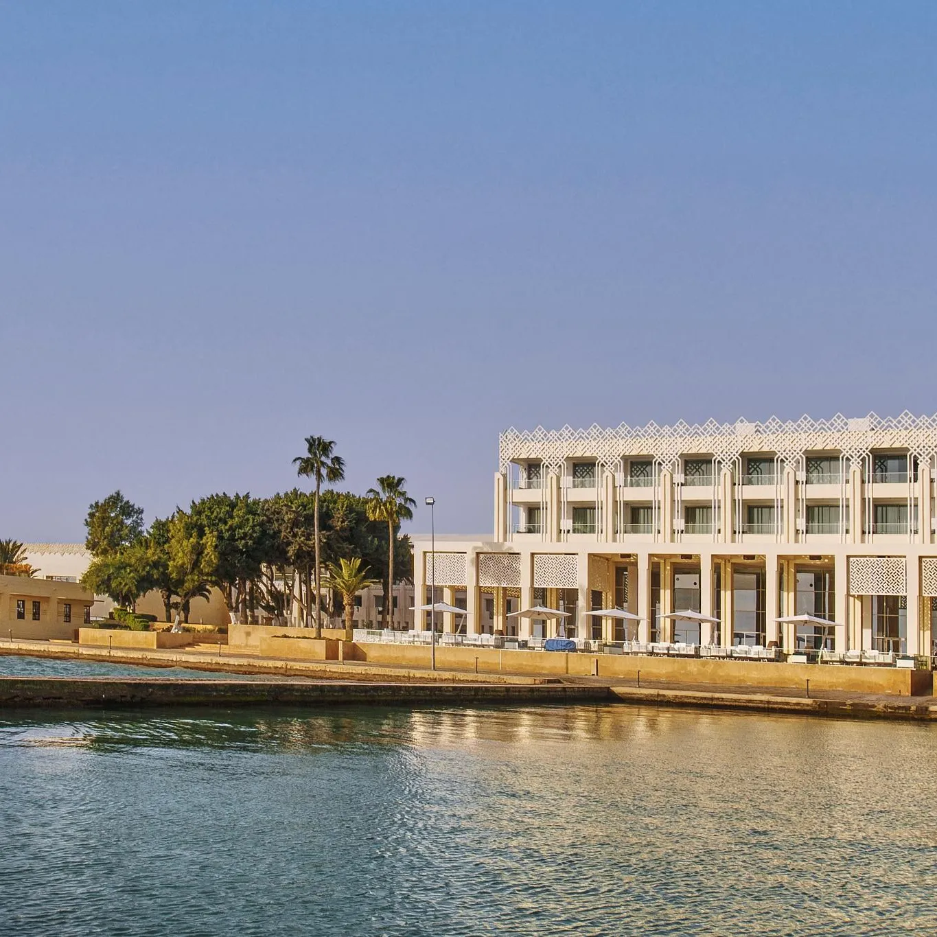 Un bâtiment moderne avec des arches et des balcons surplombe une étendue d'eau calme, entouré de palmiers et d'arbres