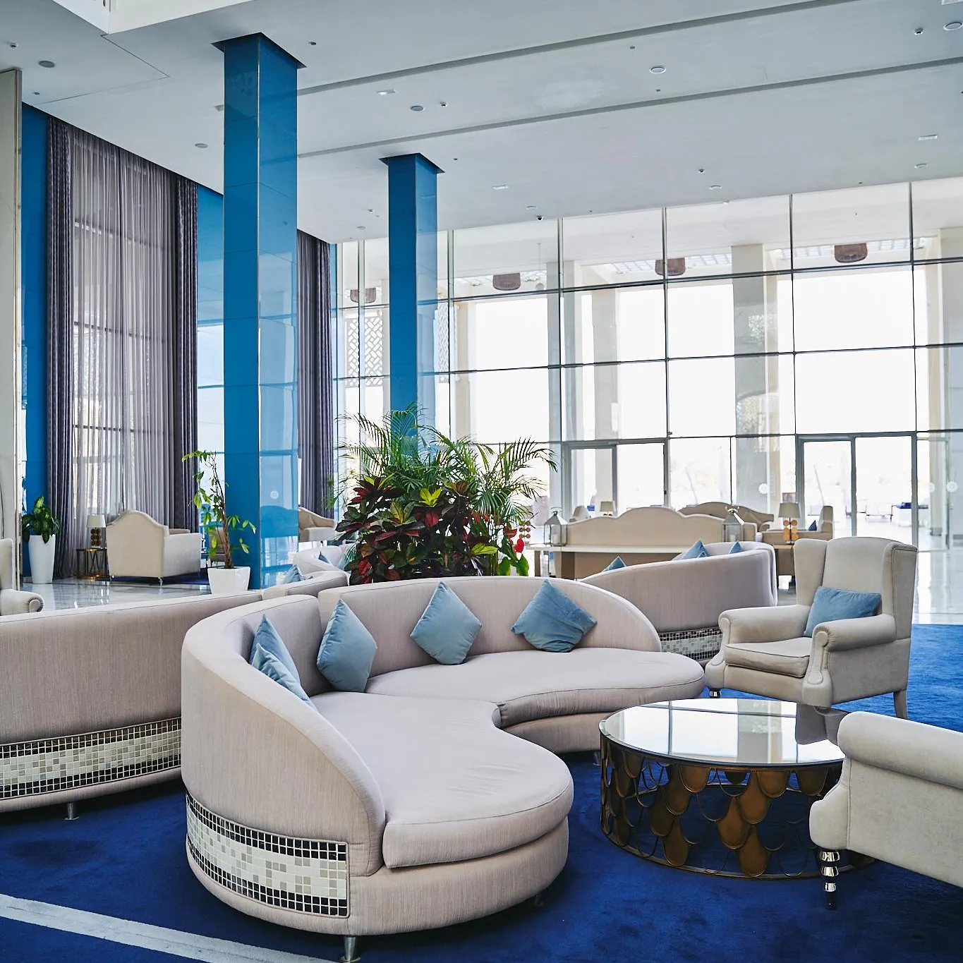 Un salon d'hôtel moderne avec des canapés et des fauteuils beiges, des coussins bleus, une table basse et des plantes vertes, devant de grandes fenêtres