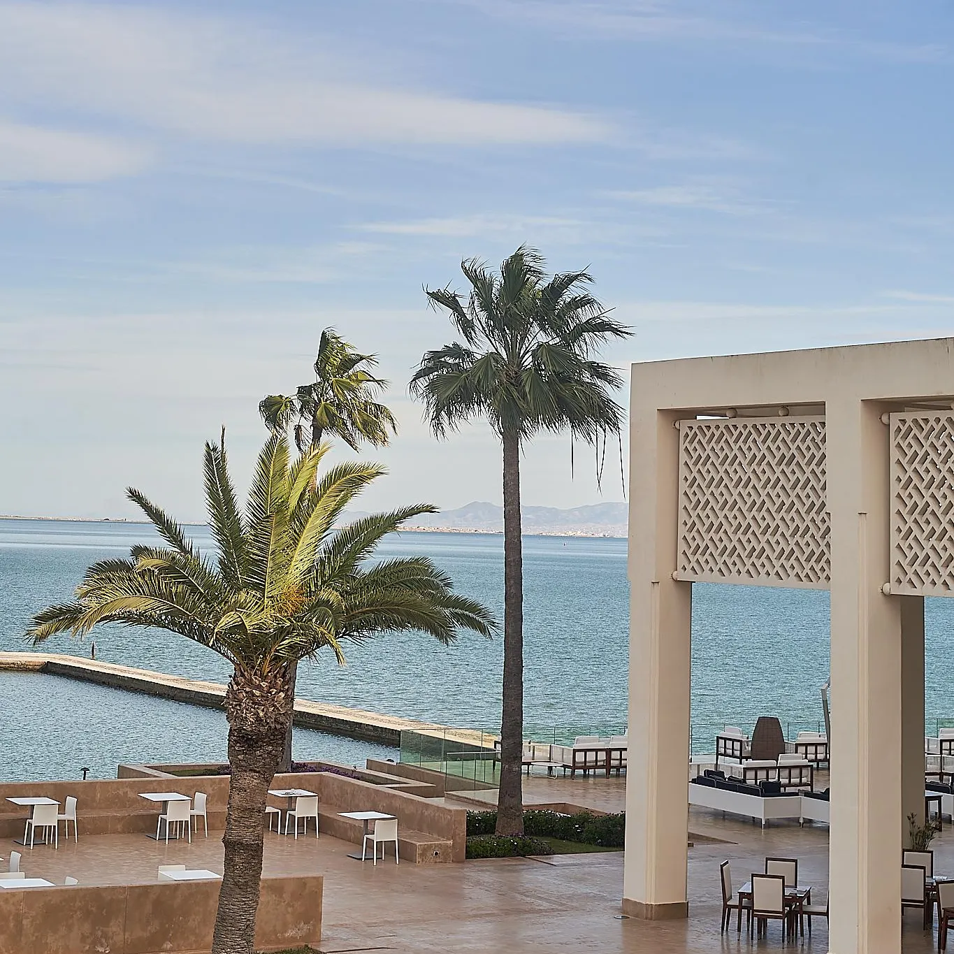 Terrasse avec tables et chaises près de la mer, bordée de palmiers et d'une structure architecturale avec des motifs géométriques