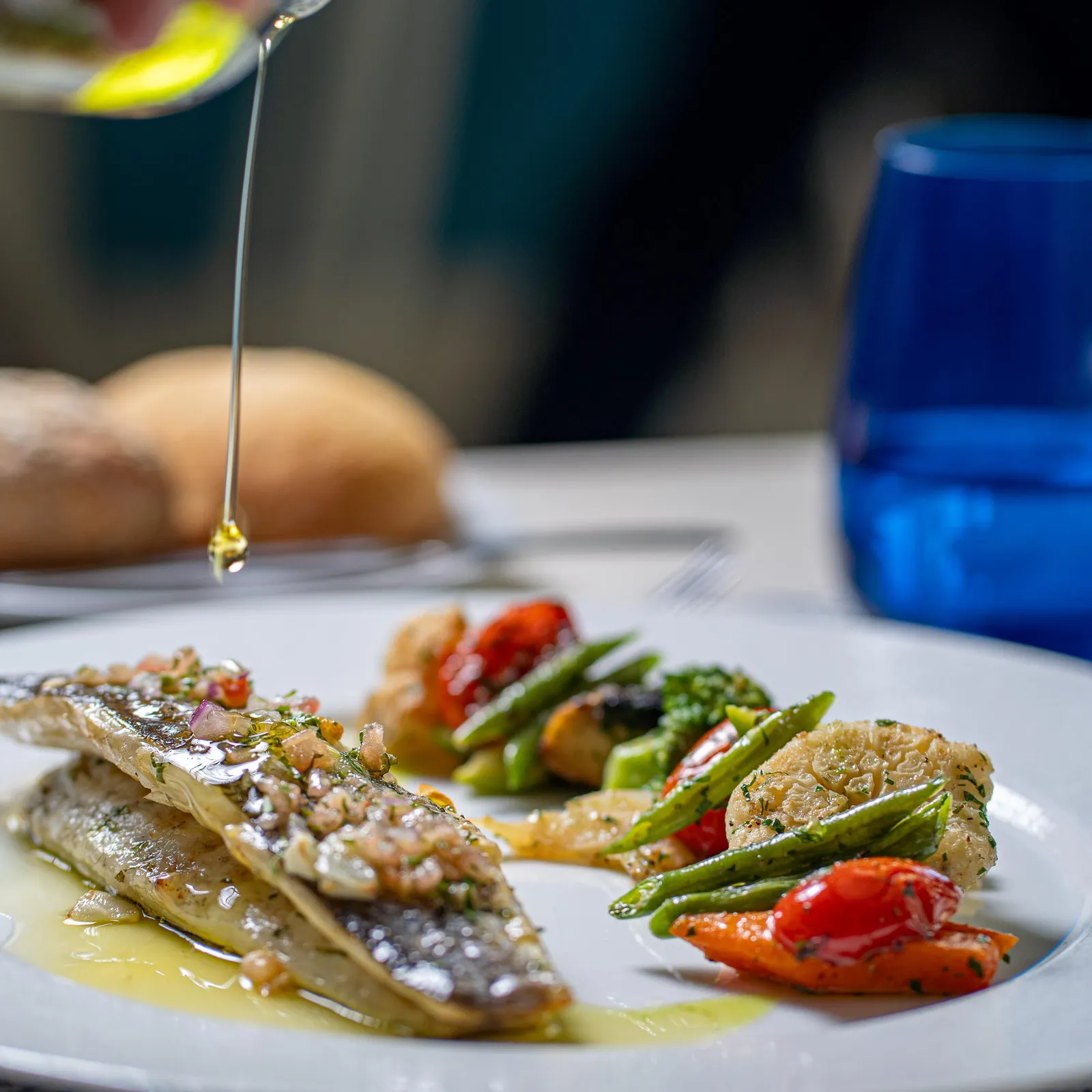 Une main verse de l'huile sur un plat composé de poisson et de légumes grillés, accompagné de pain