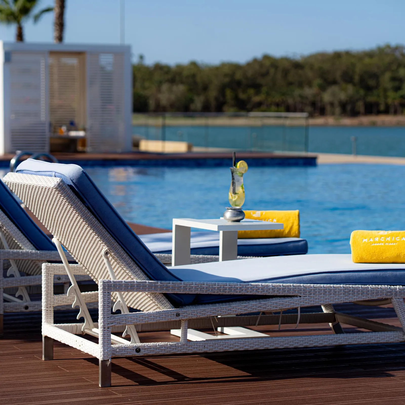 Deux transats en rotin avec coussins bleus et une serviette jaune sont disposés près d'une piscine, avec une table basse et un verre contenant une boisson