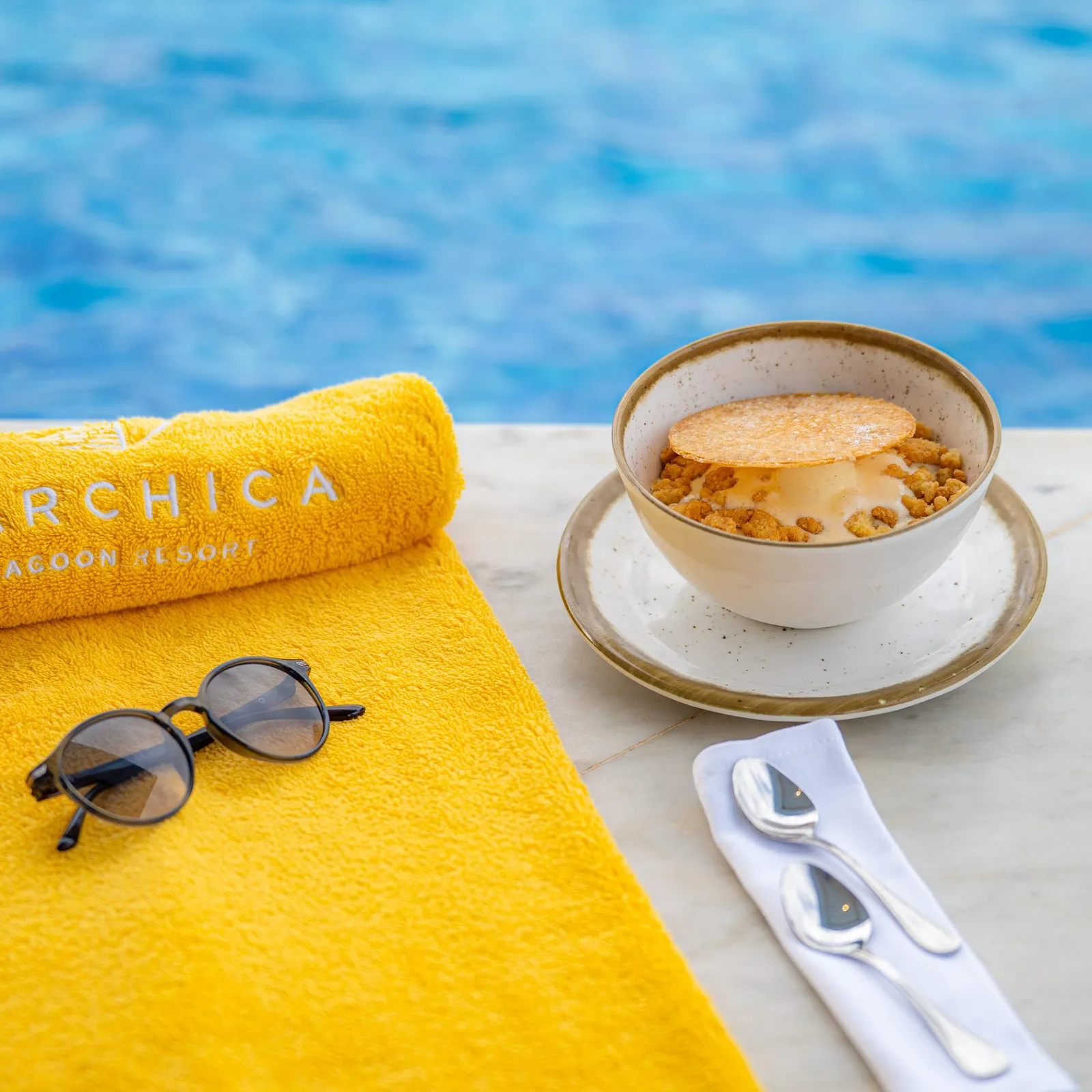 Un verre de dessert avec une couche croustillante, une serviette jaune, des lunettes de soleil et une cuillère sur une table près d'une piscine