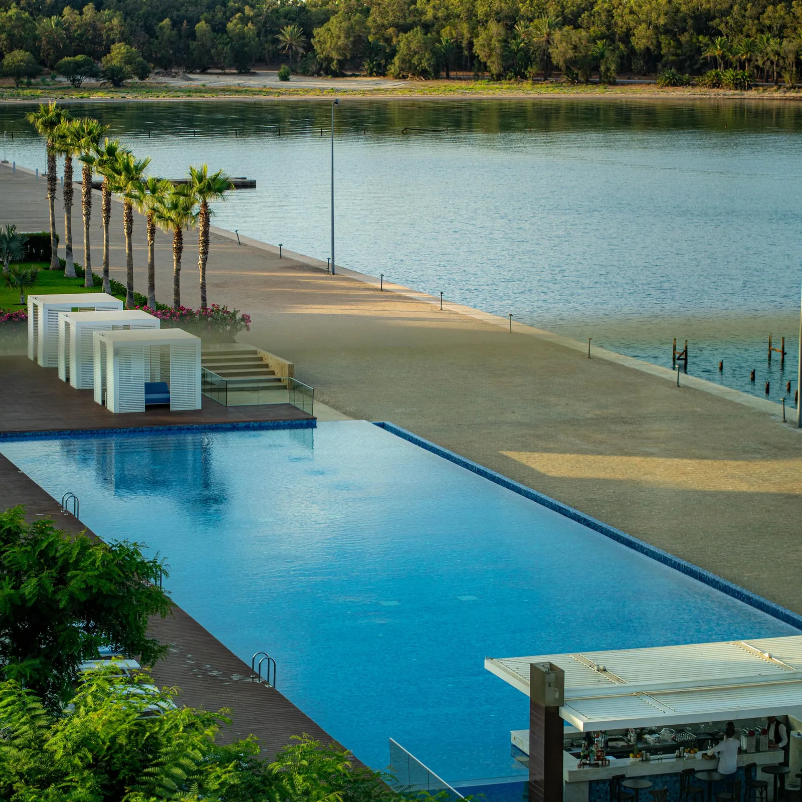 Piscine rectangulaire avec un bar et des cabanes blanches, bordée de palmiers, face à un lac et une plage de sable