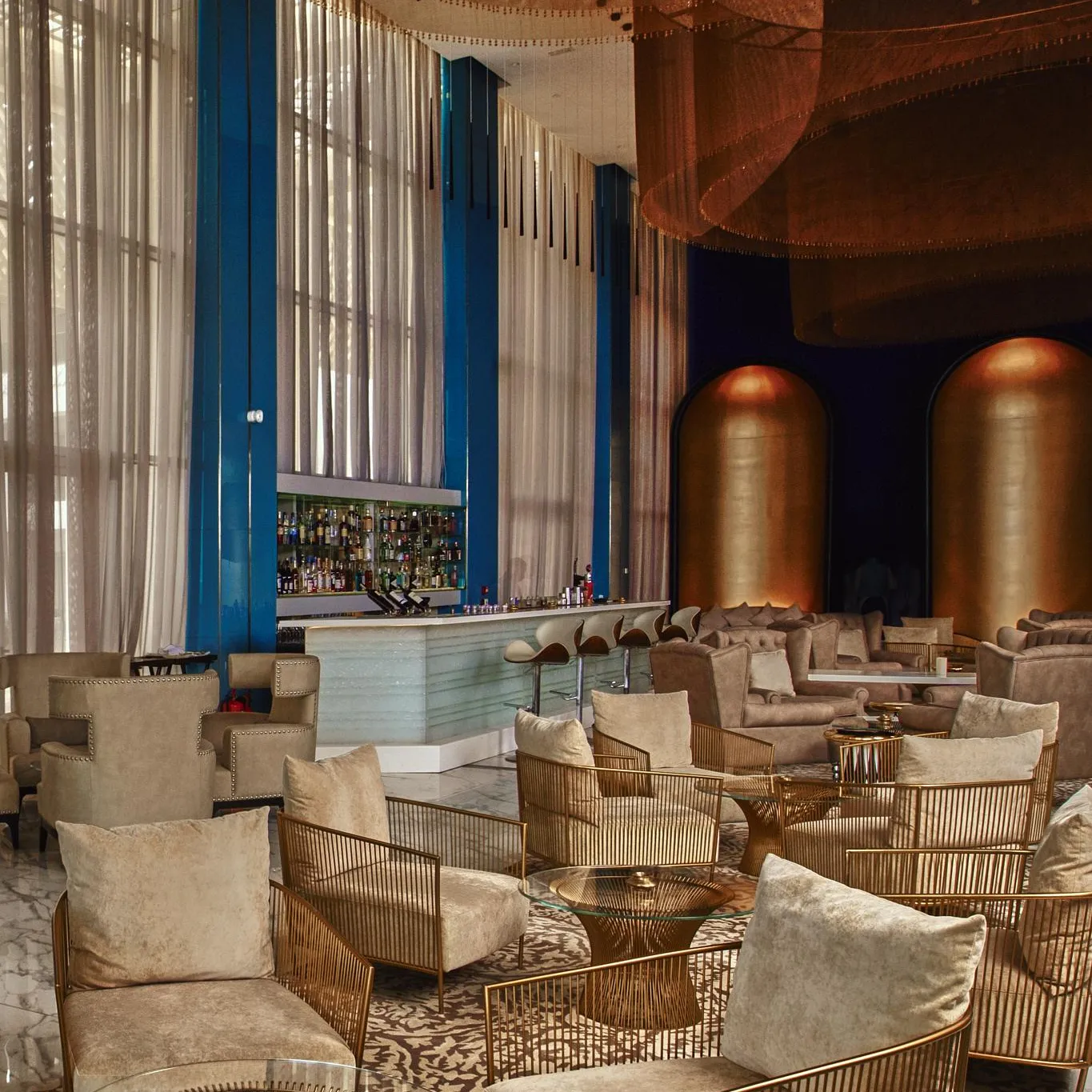 Un salon élégant avec des fauteuils en velours beige, des tables basses en verre et métal, un bar bien éclairé et des rideaux transparents devant de grandes fenêtres