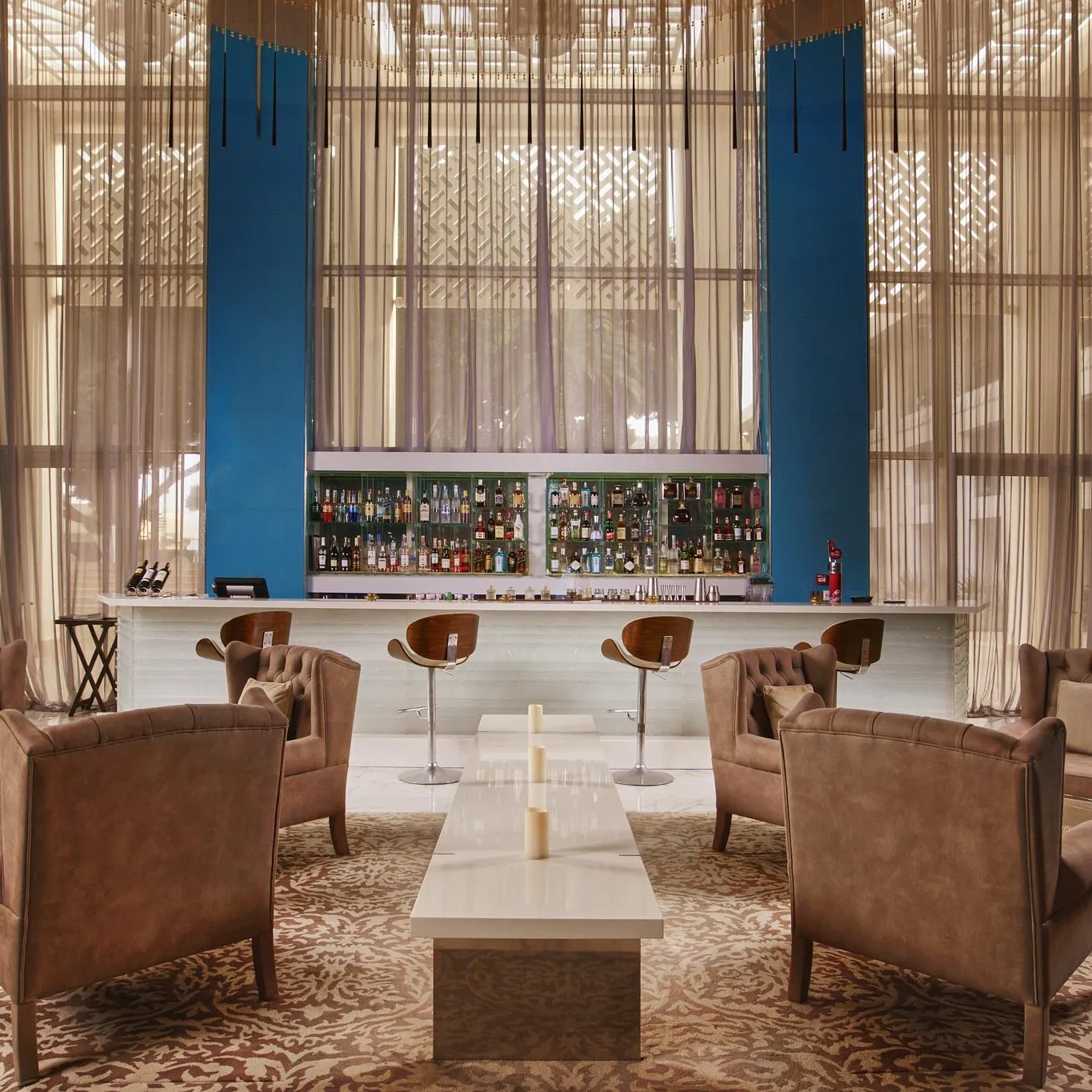 Un salon avec des fauteuils en cuir autour d'une table basse, face à un bar avec des étagères remplies de bouteilles, derrière de grandes fenêtres aux rideaux transparents