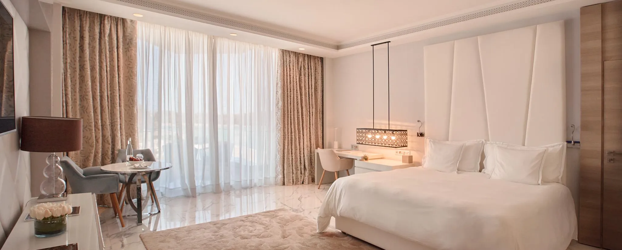 Chambre d'hôtel avec un grand lit blanc, une table de chevet avec une lampe, un coin salon près d'une grande fenêtre aux rideaux ouverts
