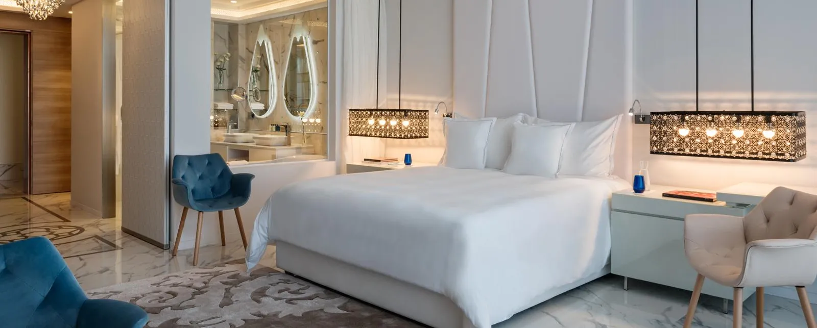 Chambre luxueuse avec lit blanc, deux fauteuils, table de chevet et vue sur une salle de bain avec miroirs ovales