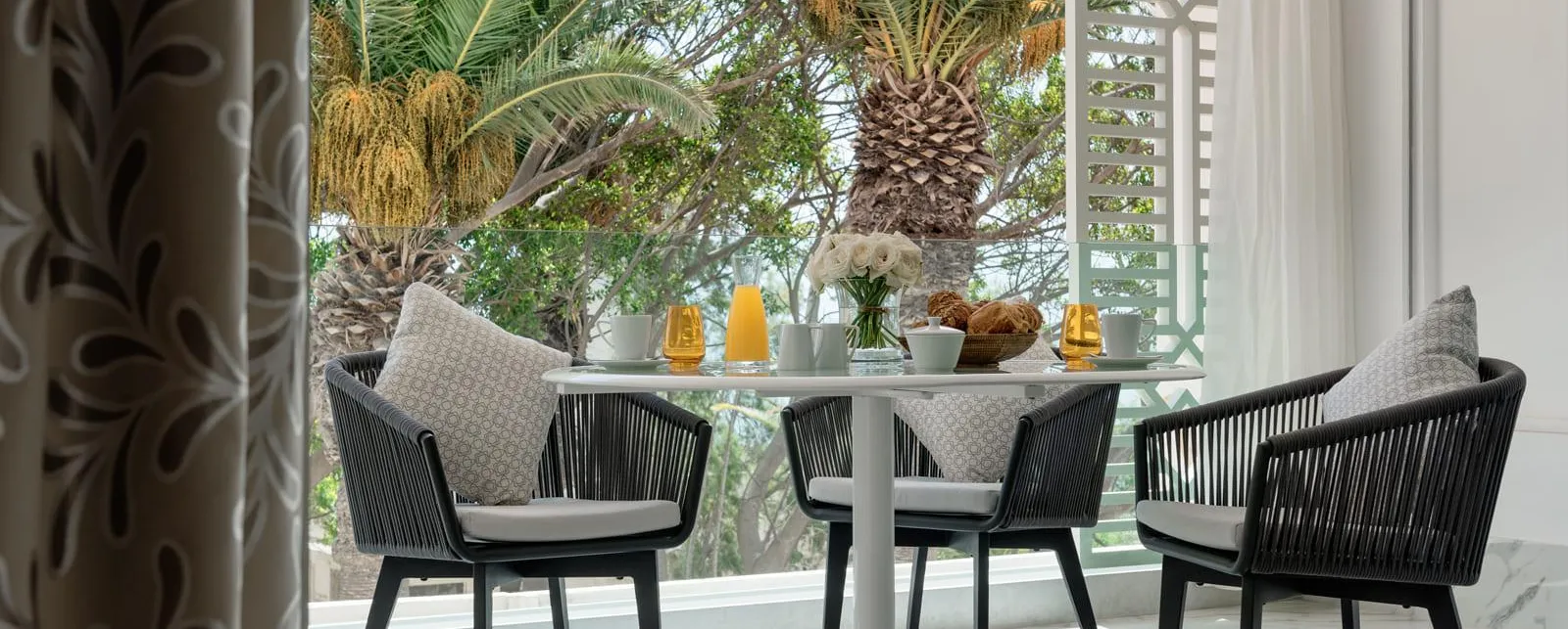 Table ronde avec quatre chaises en rotin noir et coussins gris, vue depuis une porte ouverte, avec des plantes et des éléments décoratifs sur la table en arrière-plan