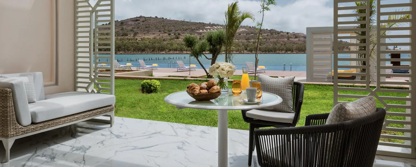 Terrasse avec table, chaises et canapé donnant sur une pelouse et un lac avec des transats et des montagnes en arrière-plan