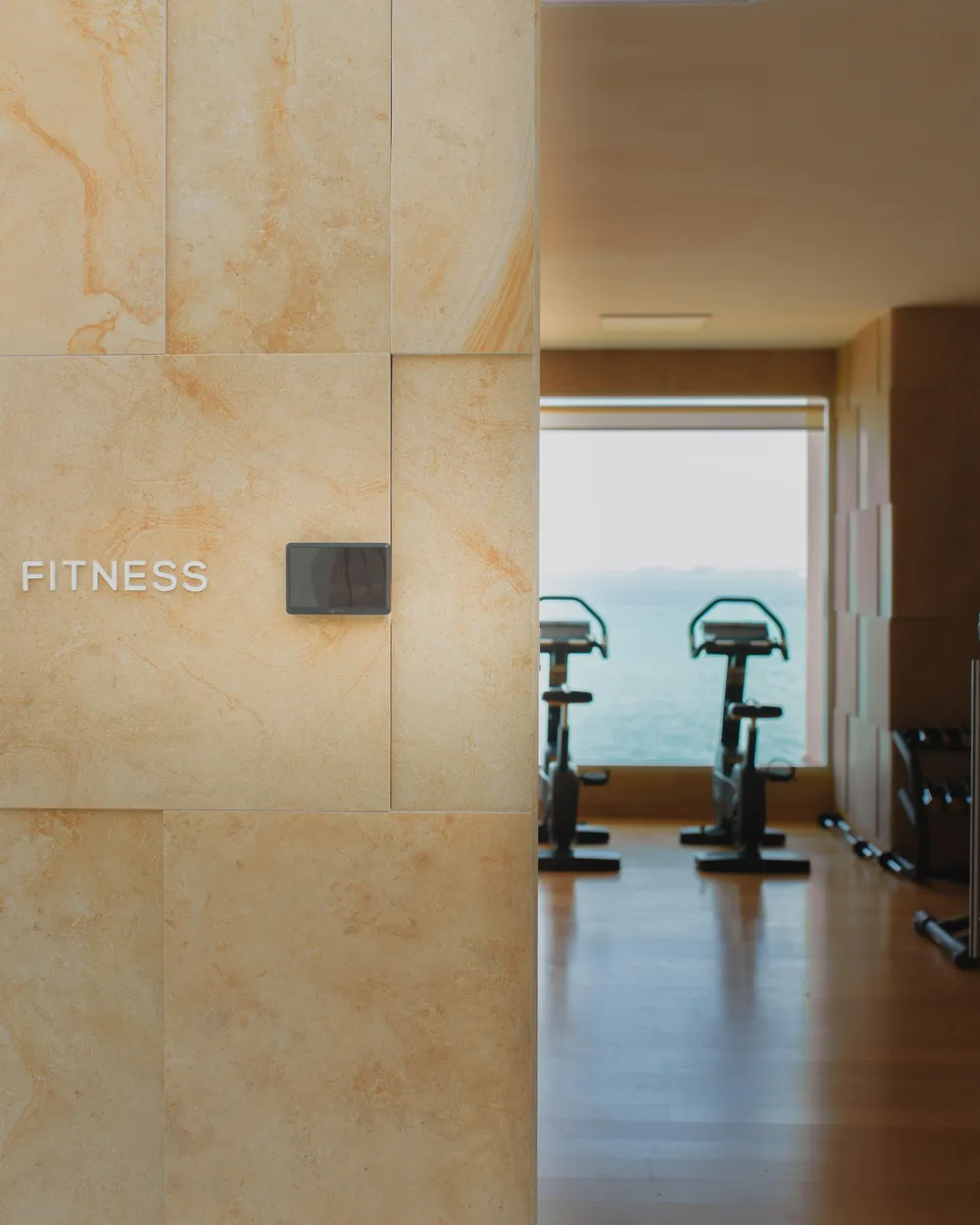 Vue d'un espace de fitness avec des vélos d'exercice, visible à travers une porte ouverte, et un mur en pierre avec l'inscription "FITNESS"