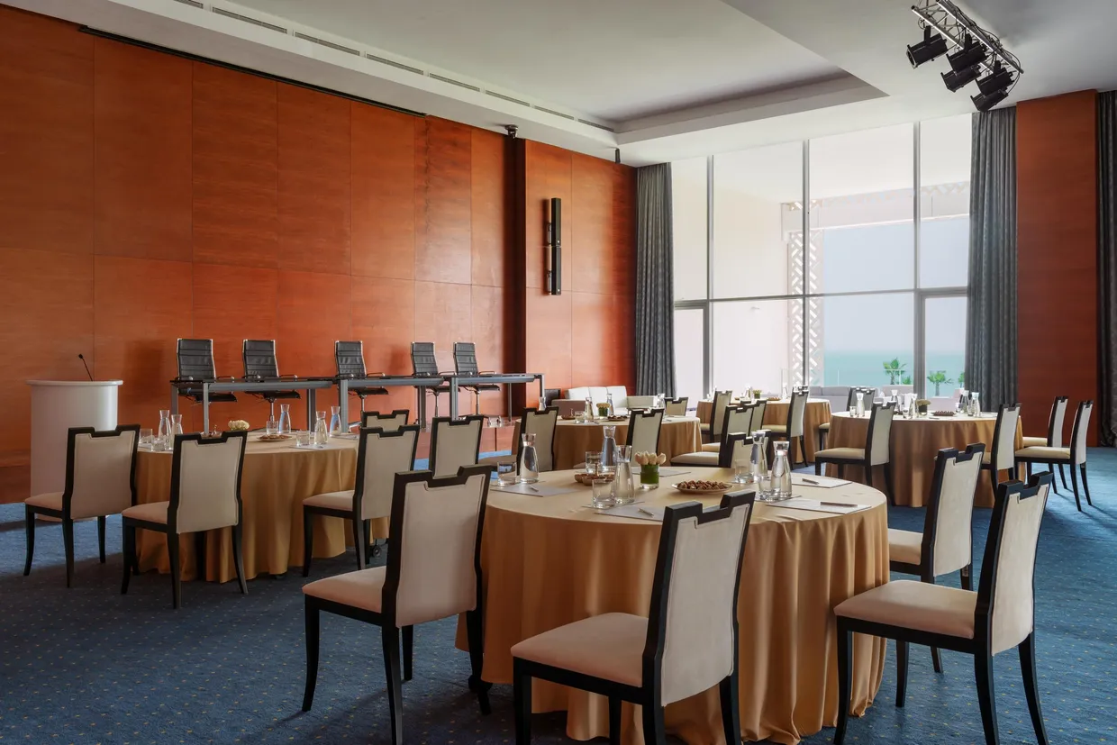 Salle de réunion avec plusieurs tables rondes recouvertes de nappes beiges, des chaises autour et une grande fenêtre avec des rideaux