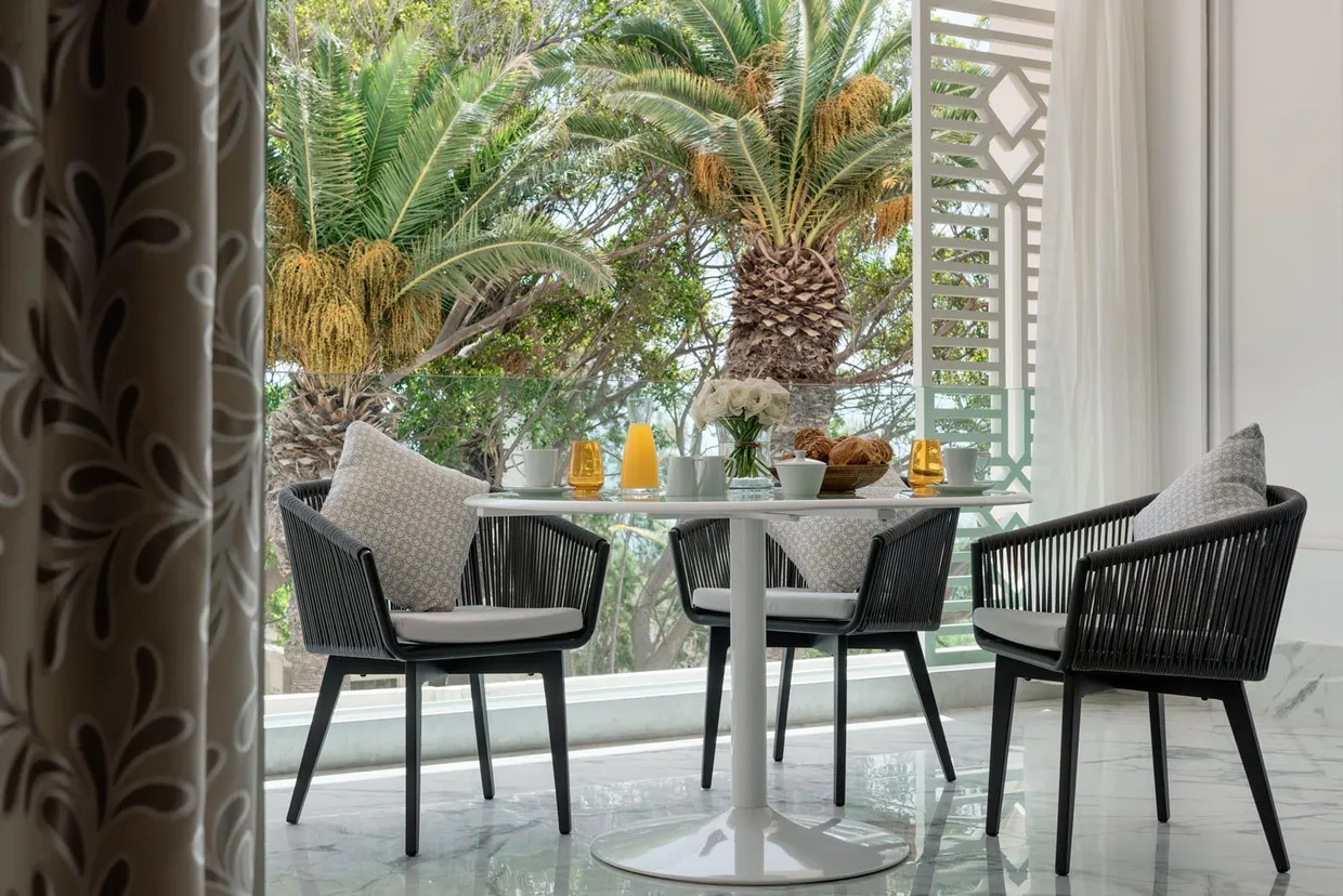 Table ronde avec quatre chaises en rotin noir et coussins gris, vue depuis une porte ouverte, avec des plantes et des éléments décoratifs sur la table en arrière-plan