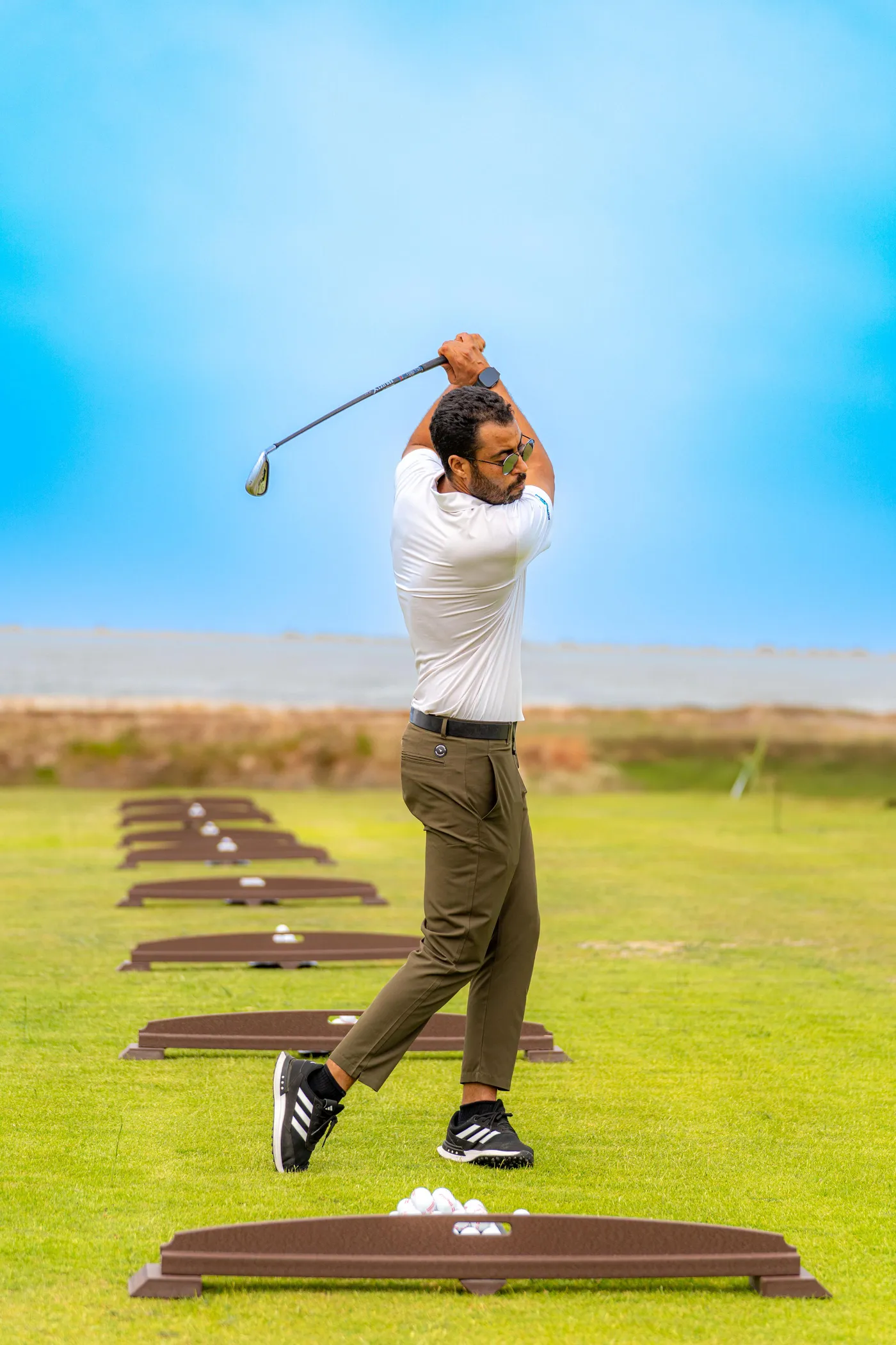 Un homme en polo blanc et pantalon vert frappe des balles de golf sur un practice en extérieur