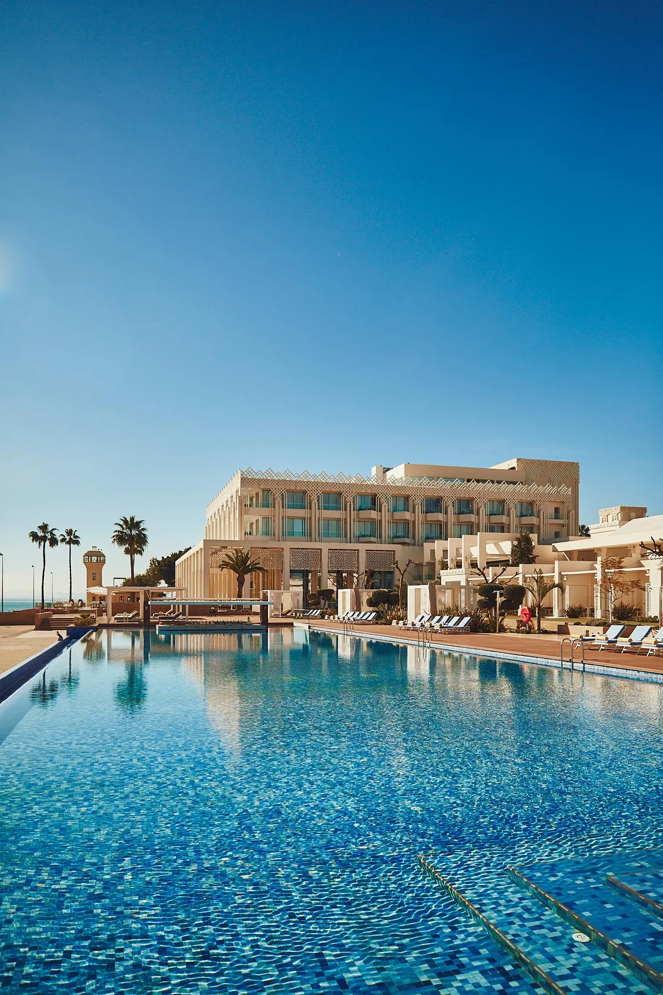 Piscine rectangulaire avec transats et parasols devant un hôtel de plusieurs étages sous un ciel bleu clair
