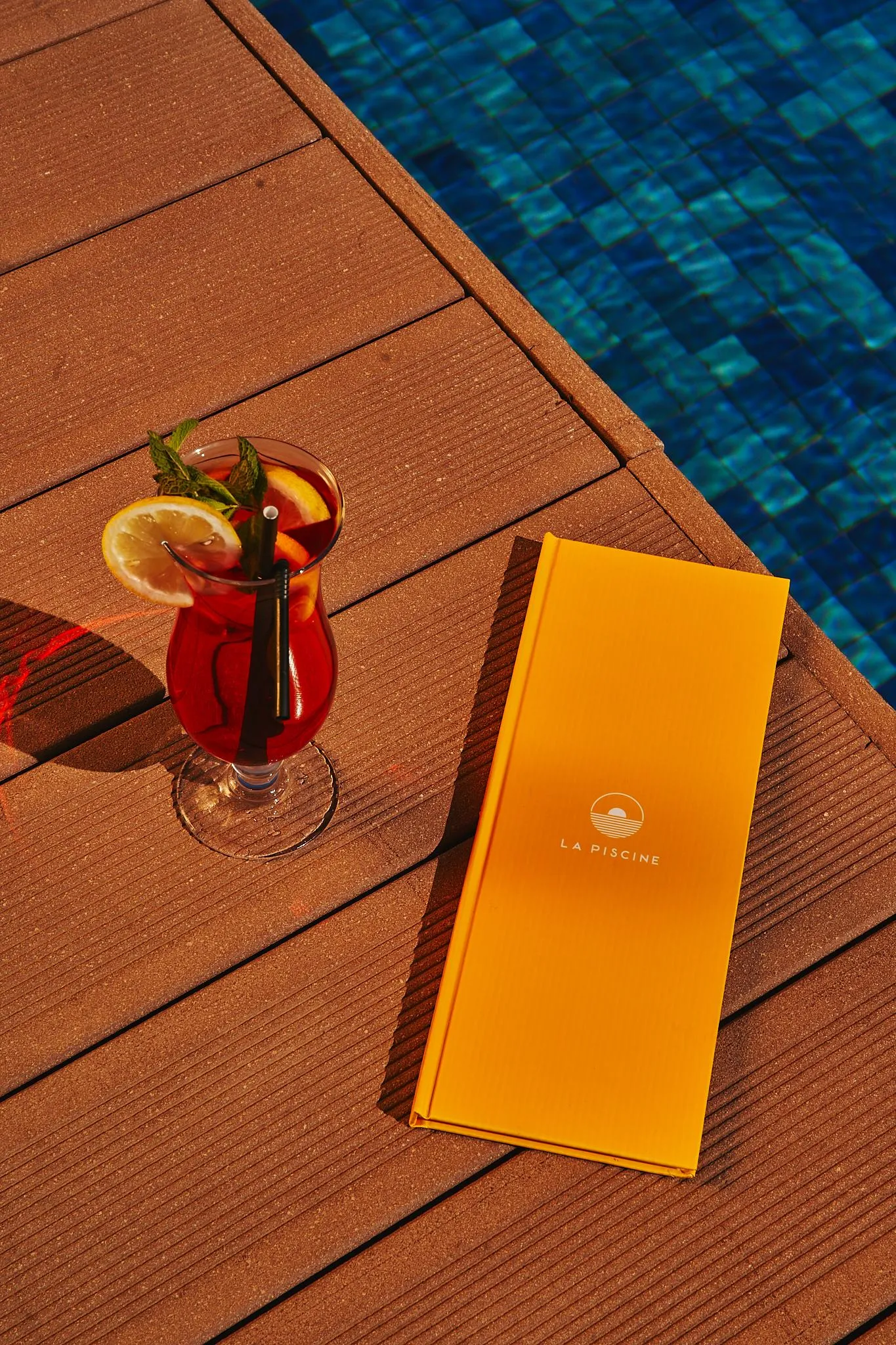 Un verre de boisson rouge avec une tranche d'orange et une paille, un menu orange et une piscine bleue en arrière-plan