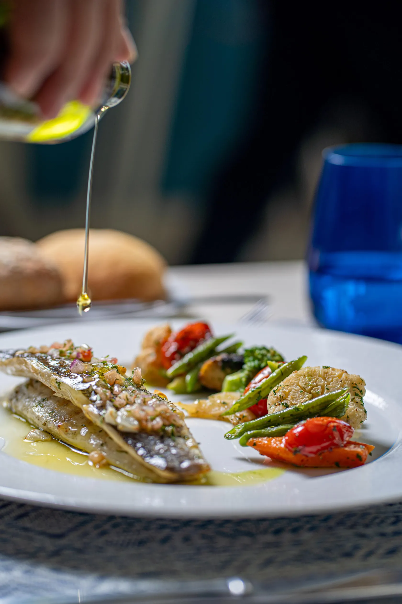 Une main verse de l'huile sur un plat composé de poisson et de légumes grillés, accompagné de pain