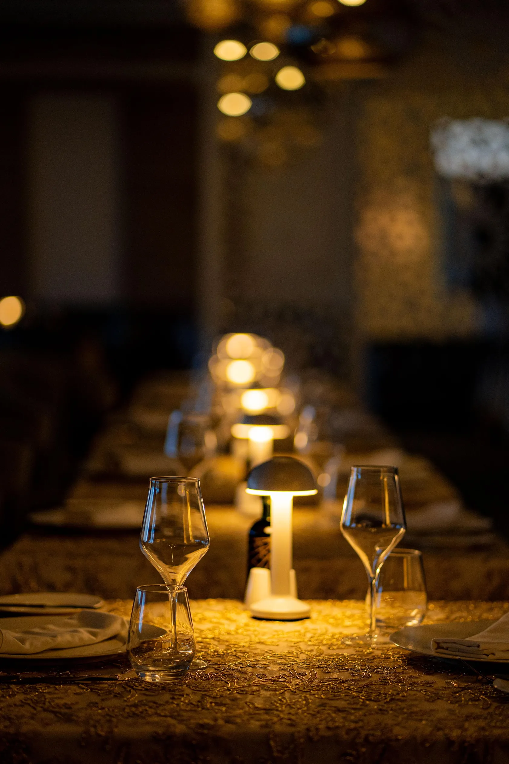 Table dressée avec verres à vin, nappe et bougies, éclairée par des lampes en fond