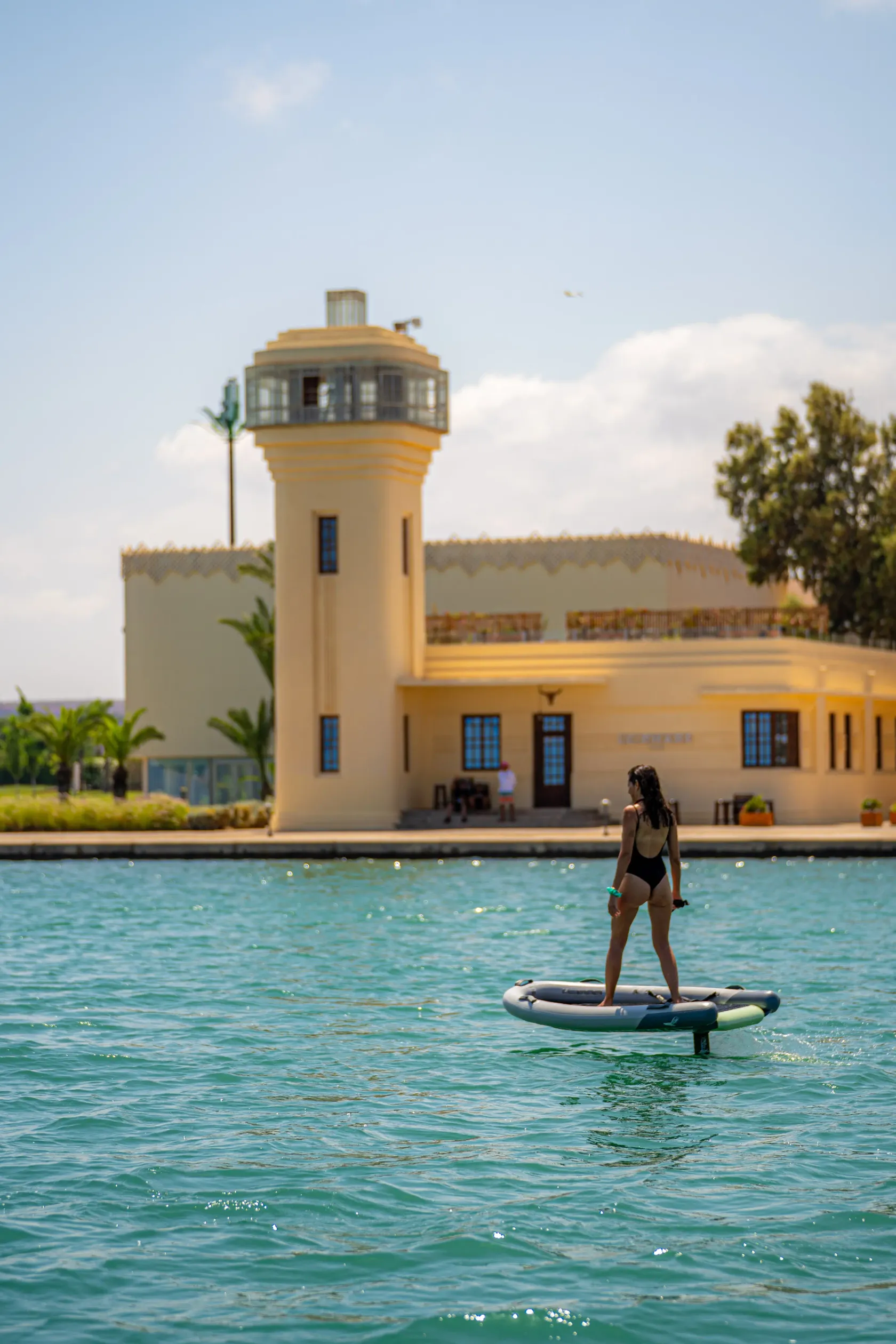Une personne en maillot de bain fait du paddle sur une étendue d'eau devant un bâtiment jaune avec une tour