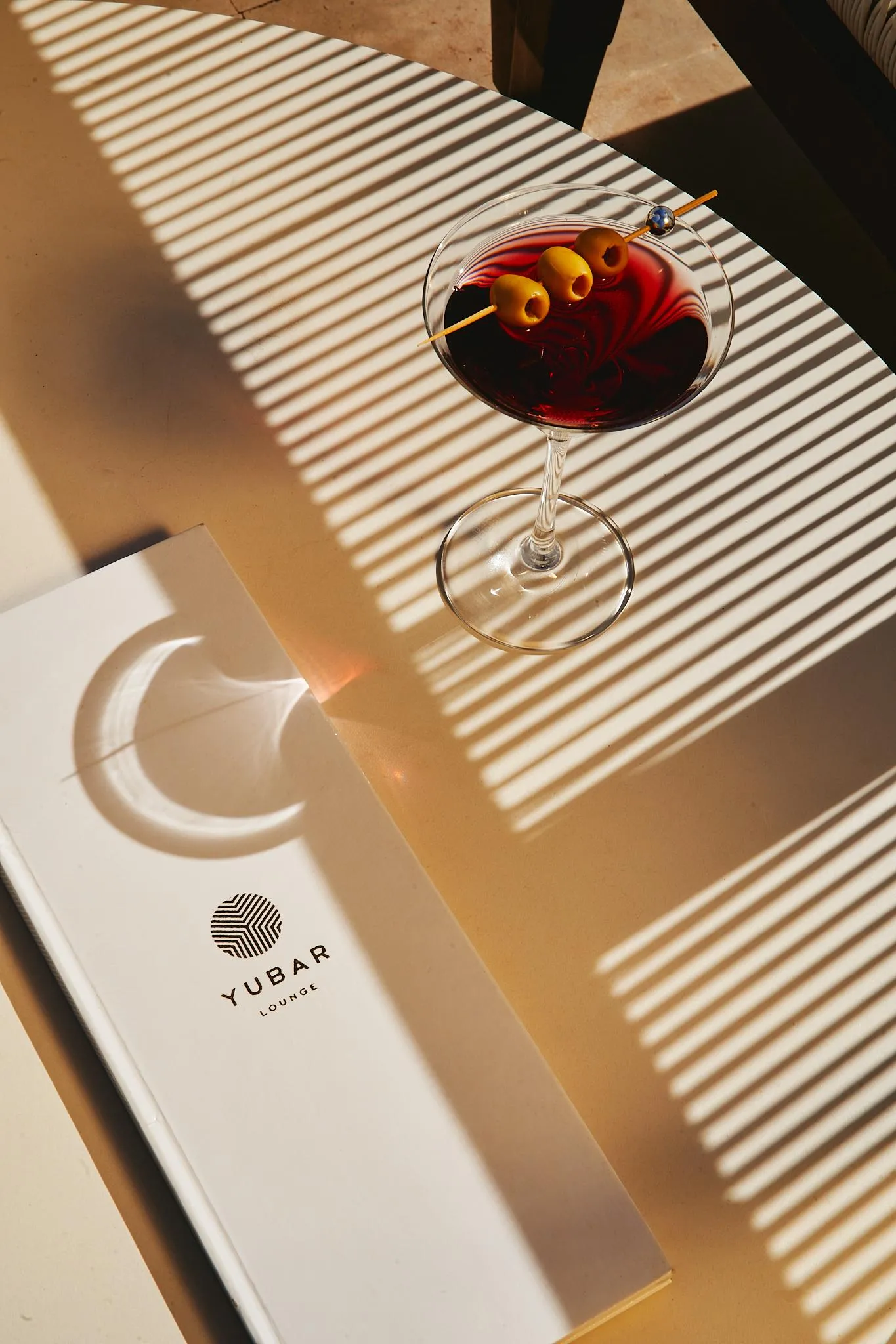 Un verre de cocktail avec des olives et une paille, posé sur une table en bois avec un menu "YUBAR LOUNGE" à côté