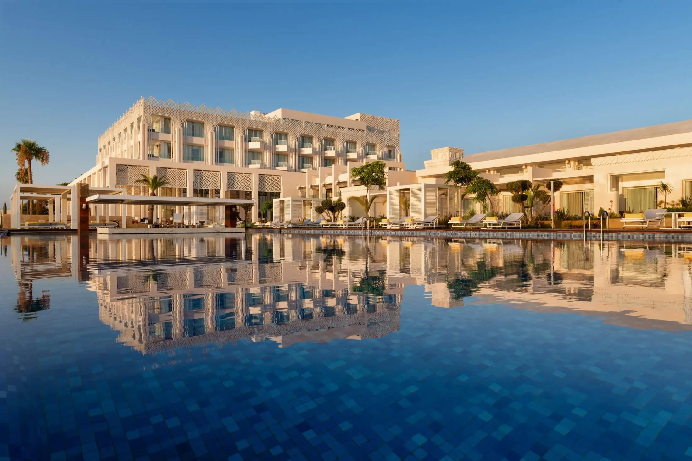 Un grand bâtiment blanc avec des balcons et des motifs géométriques, reflété dans une piscine bleue, entouré de palmiers et de transats