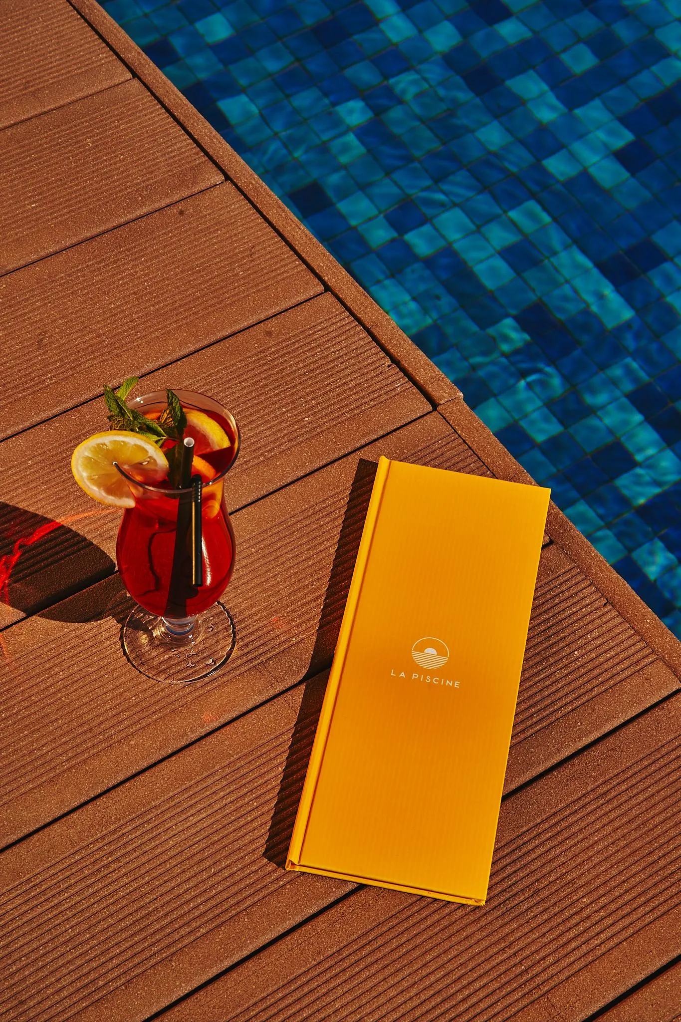 Un verre de boisson rouge avec une tranche d'orange et une paille, un menu jaune et une piscine bleue en arrière-plan