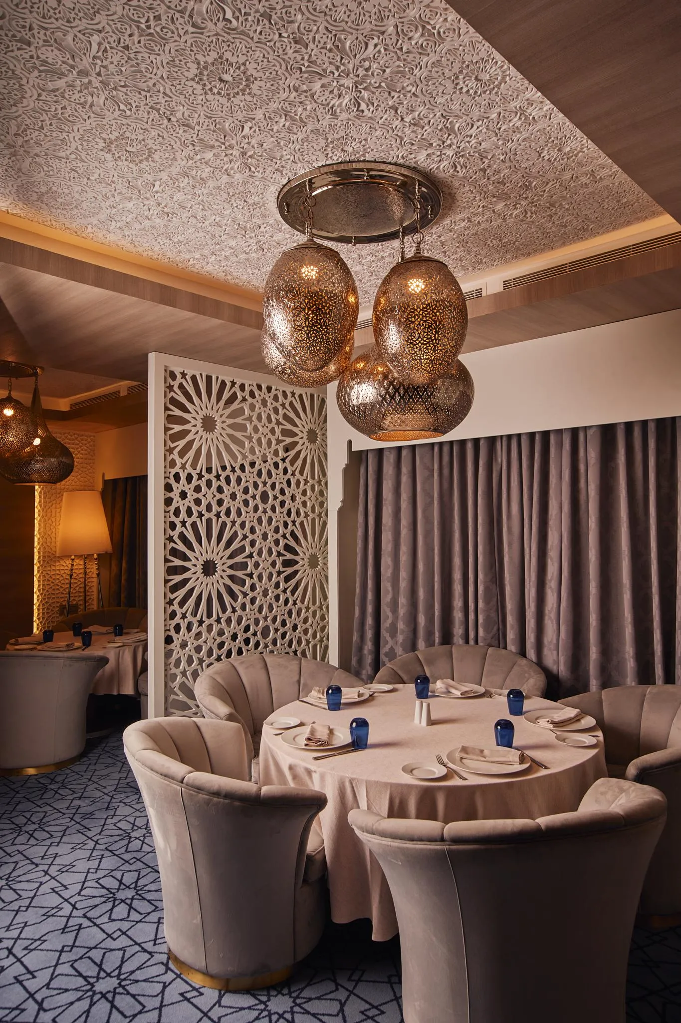 Un restaurant élégant avec des tables rondes, des chaises rembourrées en velours beige, des luminaires en métal et un plafond décoré de motifs géométriques