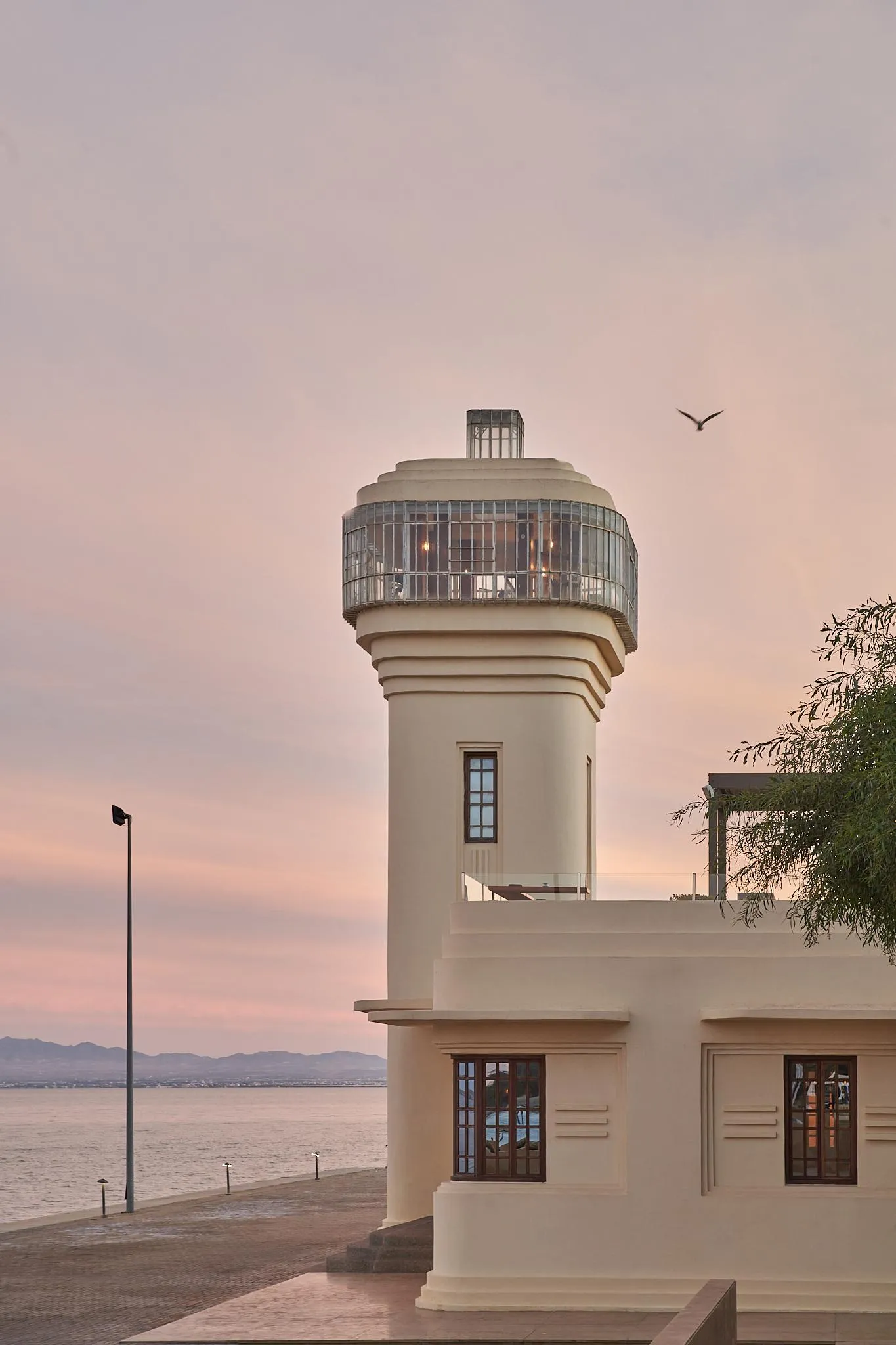 Un phare moderne avec une lanterne vitrée en haut se dresse près d'une étendue d'eau au coucher du soleil