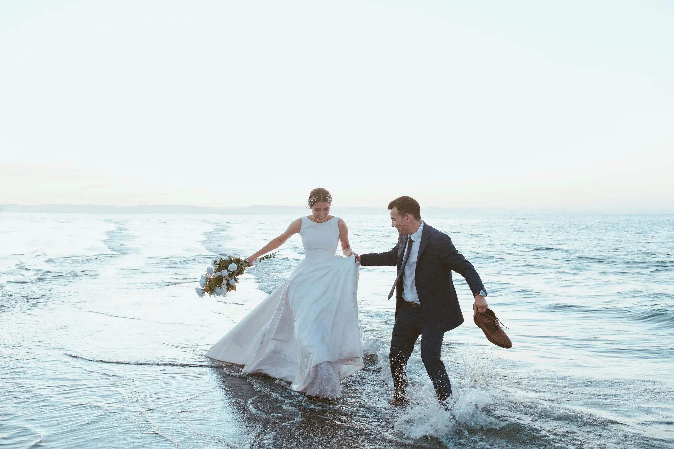 Un couple marche pieds nus dans l'eau, la mariée tenant un bouquet et l'homme tenant ses chaussures