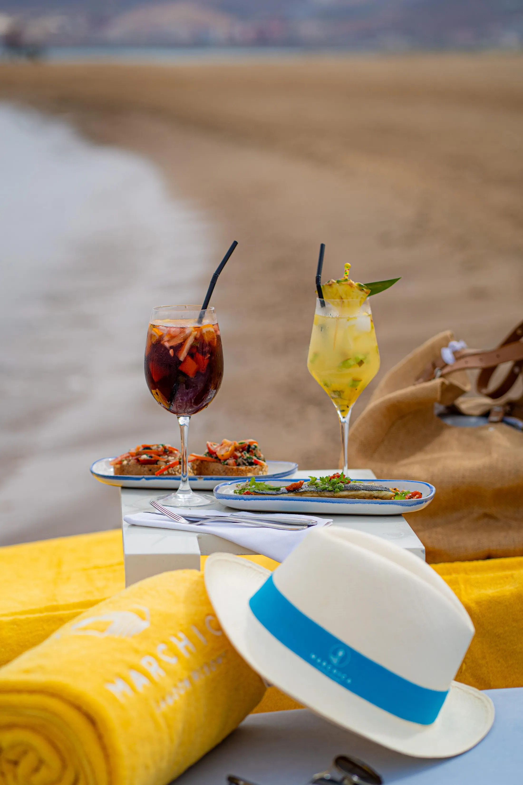 Deux verres de boissons fraîches avec des pailles, des assiettes de nourriture et un chapeau sur une table près d'une plage