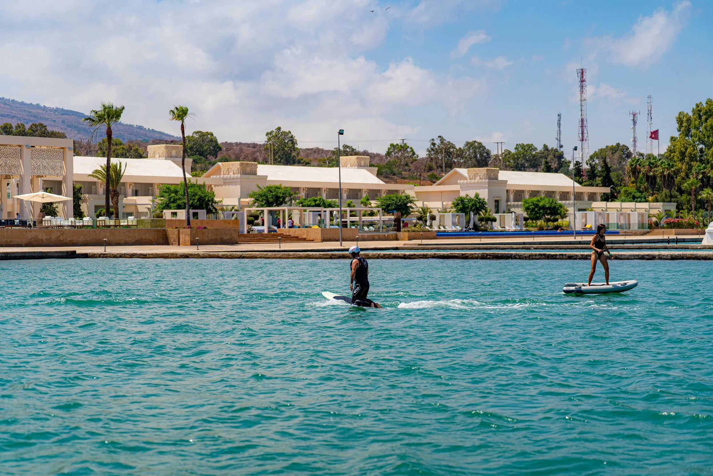 Deux personnes font du paddle sur une étendue d'eau turquoise devant un bâtiment blanc avec des palmiers