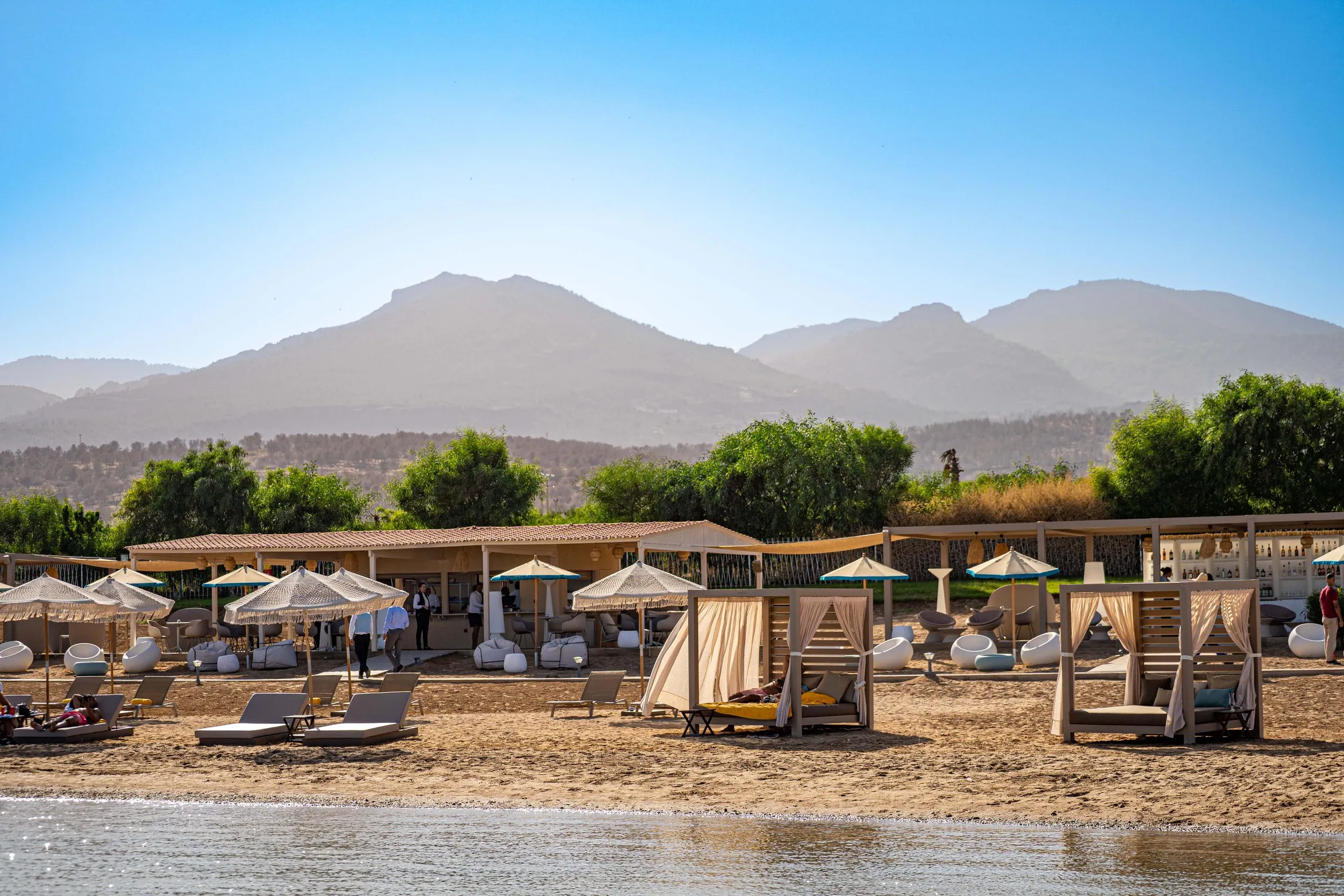 Une plage avec des transats, des parasols et des cabanes en bois, située au pied de montagnes avec une végétation dense