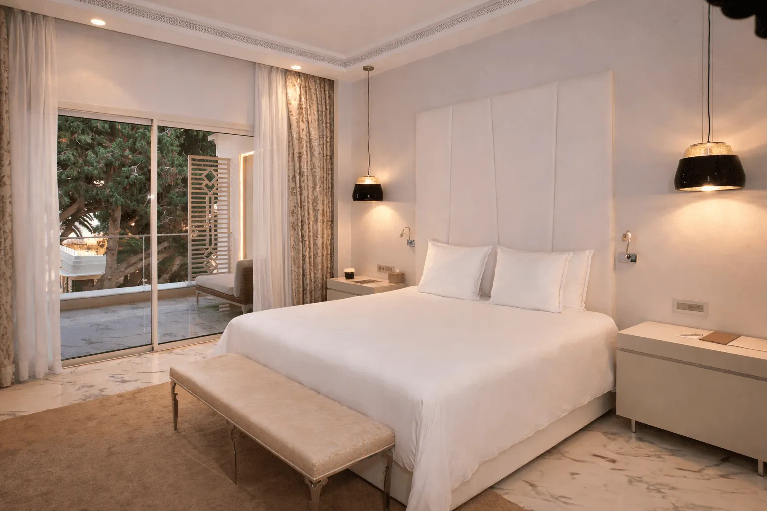 Chambre d'hôtel moderne avec un lit double, une banquette au pied du lit, des lampes suspendues et une grande baie vitrée donnant sur une terrasse avec vue sur des arbres