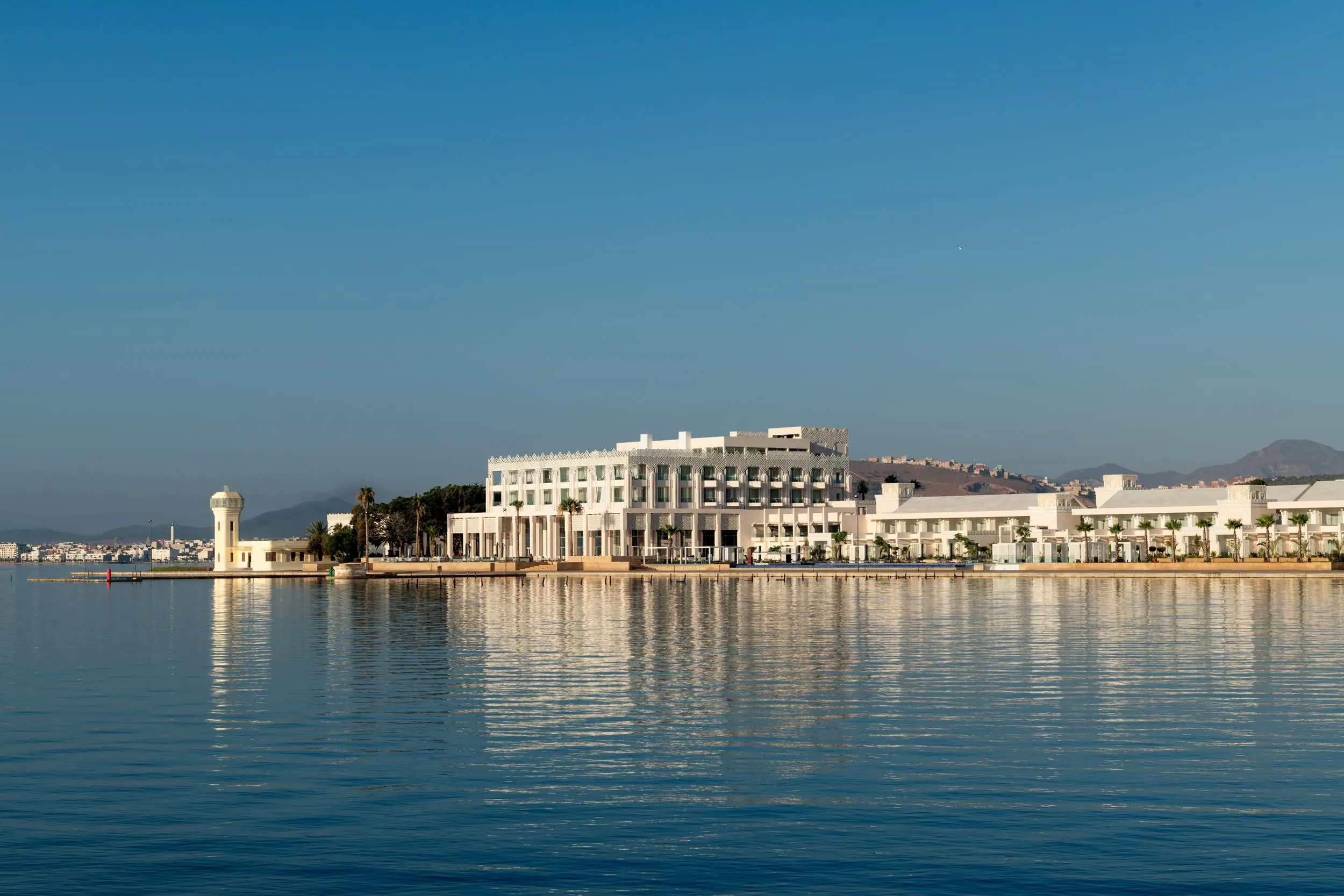 Un bâtiment blanc moderne en bord de mer avec une tour cylindrique, reflété dans l'eau calme sous un ciel bleu clair
