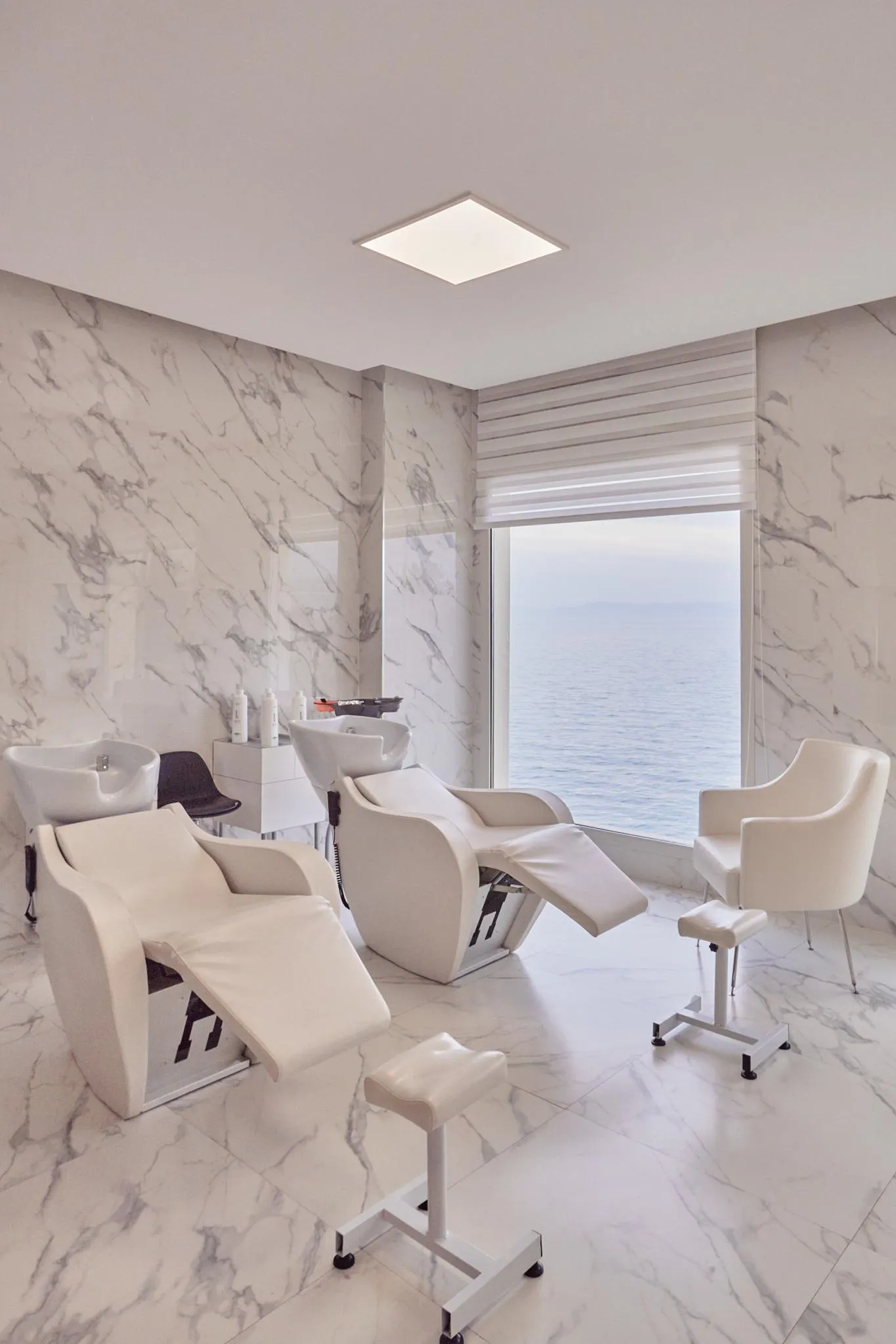 Salon de coiffure avec des fauteuils de coiffure blancs et des murs recouverts de marbre blanc, donnant sur une vue sur la mer à travers une grande fenêtre