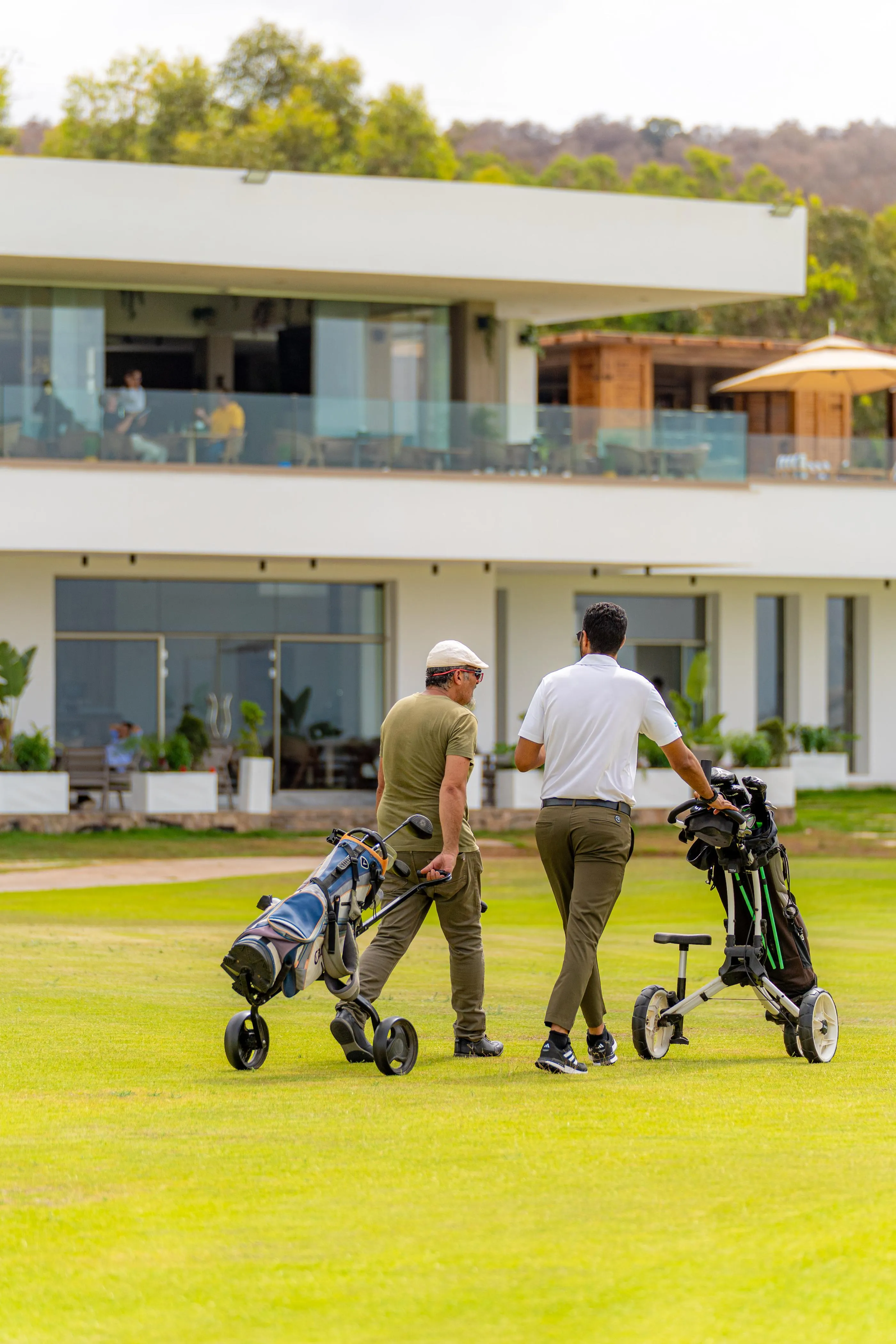 Deux hommes marchent sur un terrain de golf avec des sacs de clubs, devant un bâtiment moderne aux grandes baies vitrées