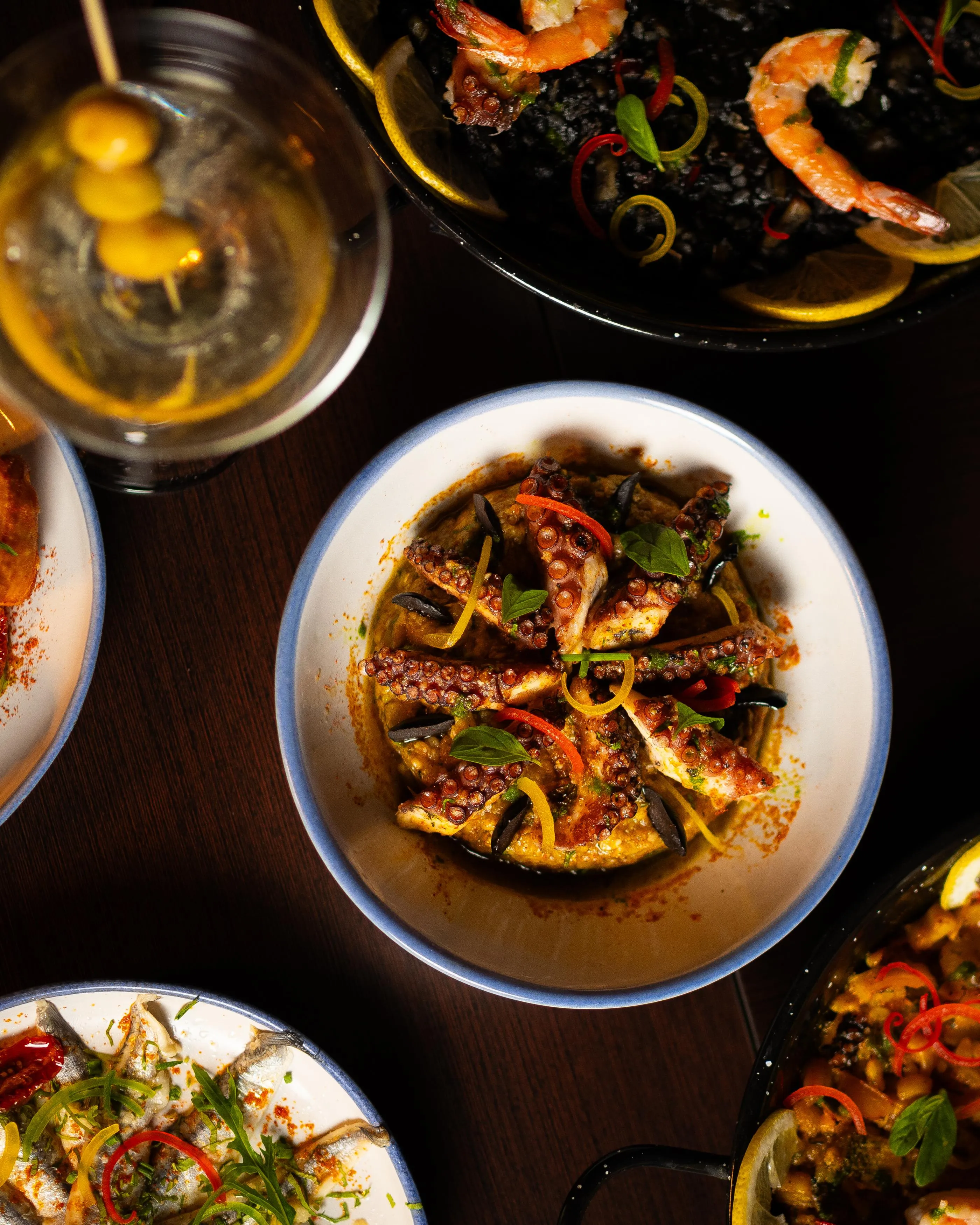 Un plat de calamars grillés avec des légumes et des herbes, accompagné d'autres assiettes de fruits de mer et d'une boisson dans un verre