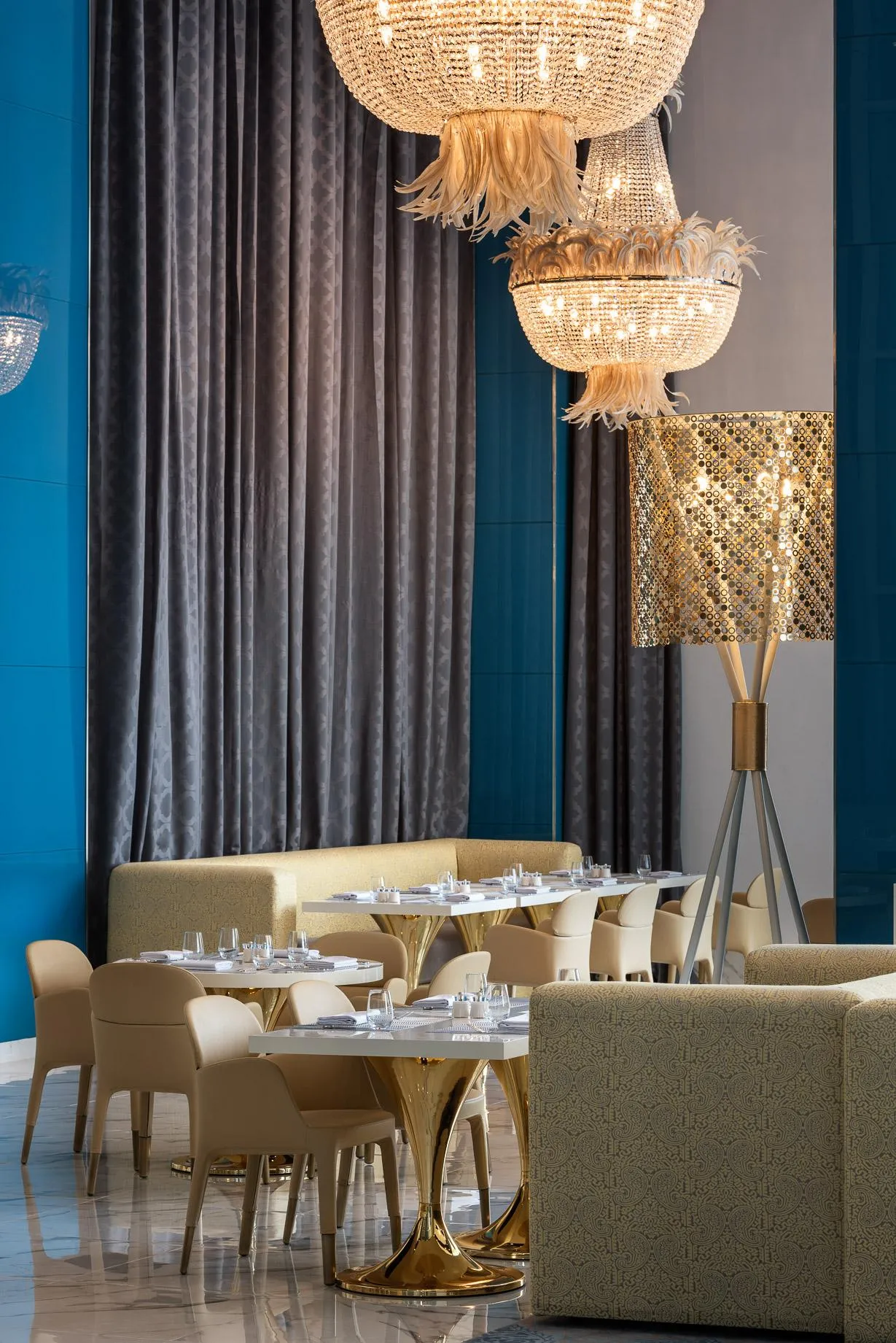 Salle de restaurant élégante avec tables rondes, chaises et canapés beiges, luminaires en cristal et rideaux gris