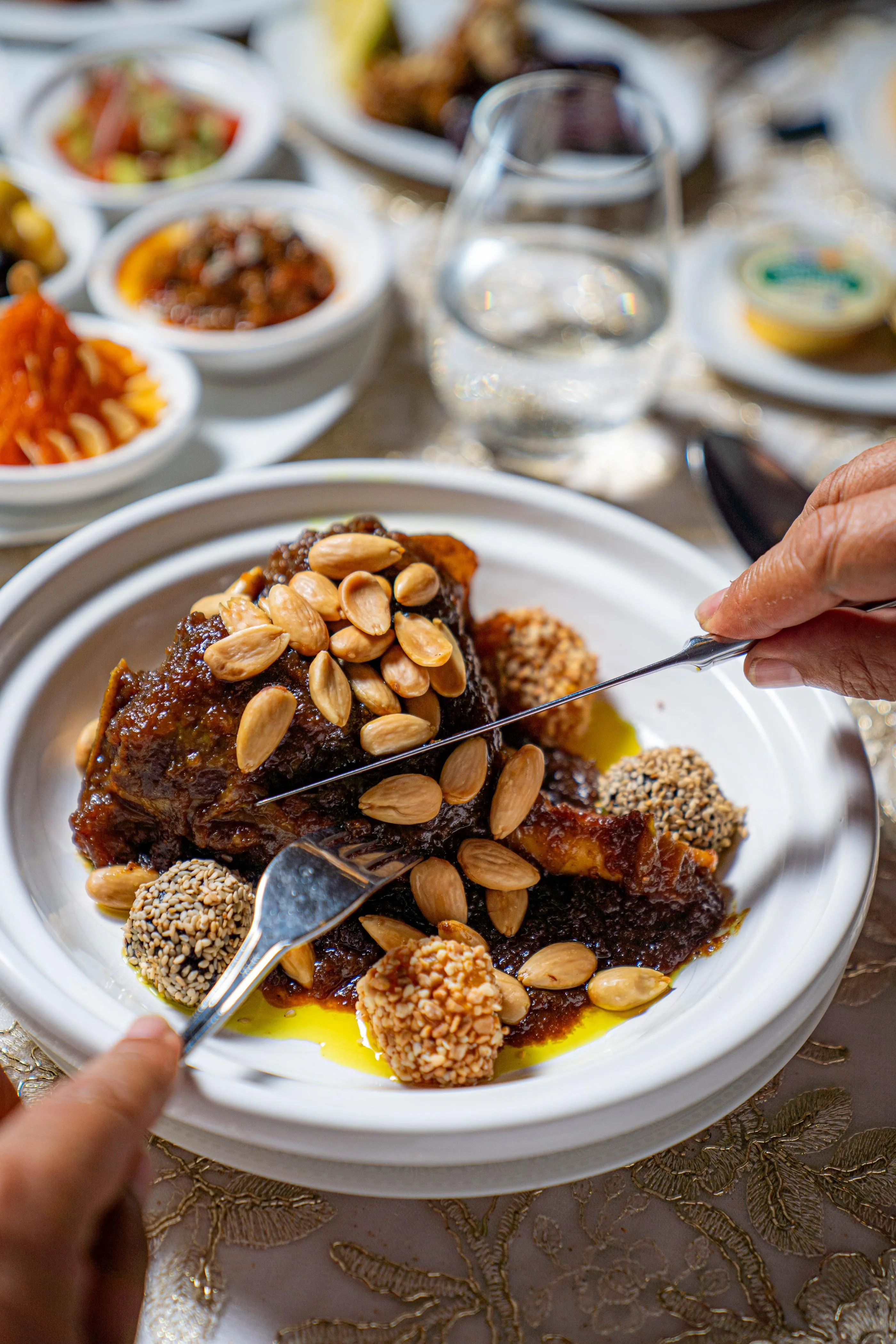 Une personne sert un plat avec des morceaux de viande, des amandes et des graines de sésame sur une table garnie de divers petits plats