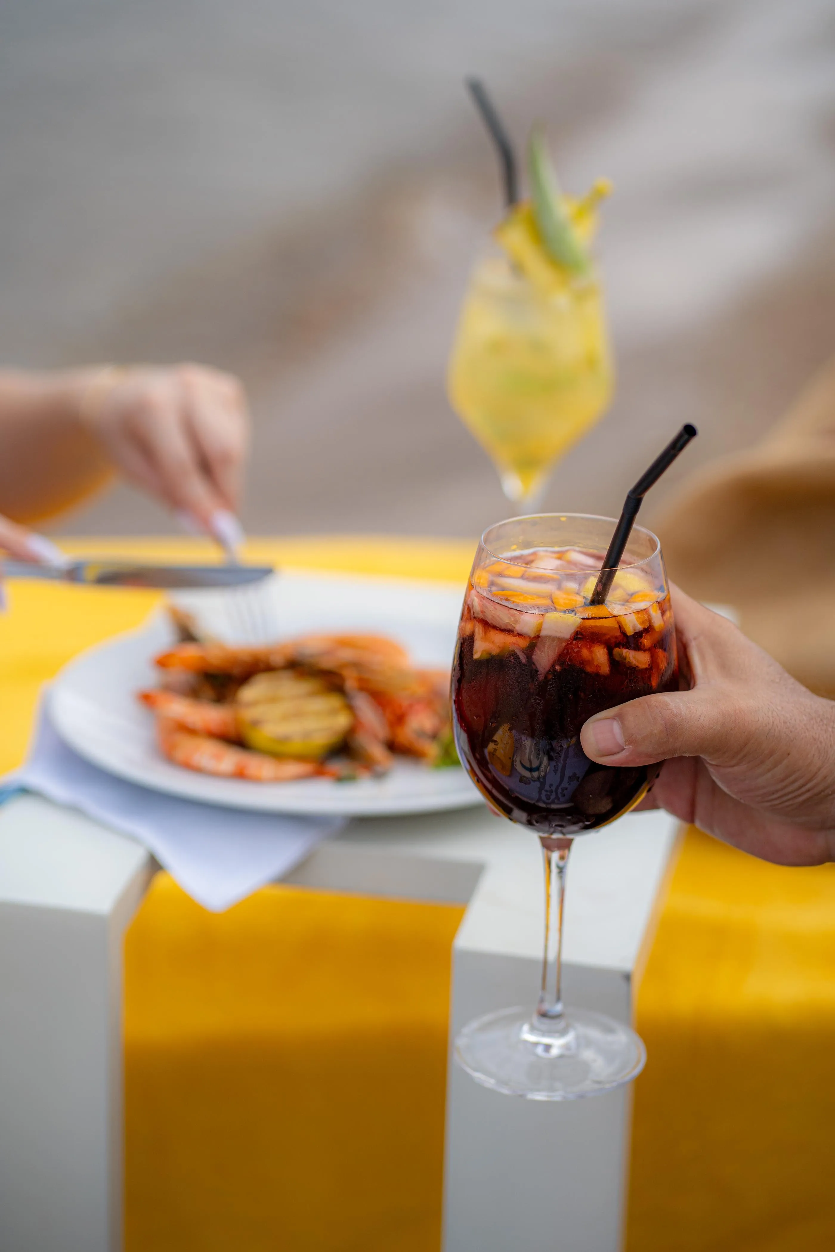 Une personne tient un verre de boisson rouge avec des glaçons et une paille, devant une assiette de nourriture et une table jaune
