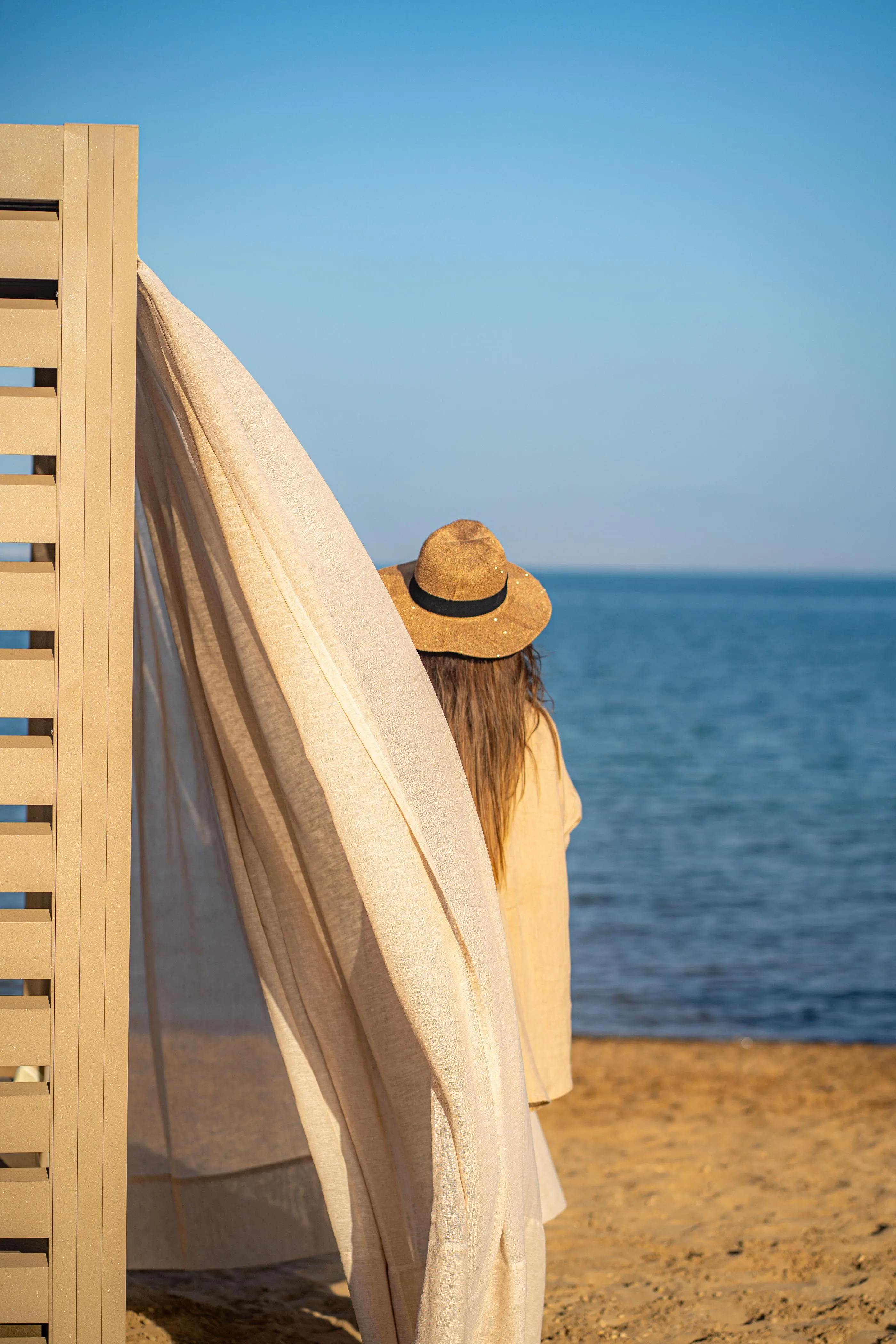 Une personne portant un chapeau regarde la mer depuis une plage, un drap blanc accroché à une structure en bois à côté d'elle