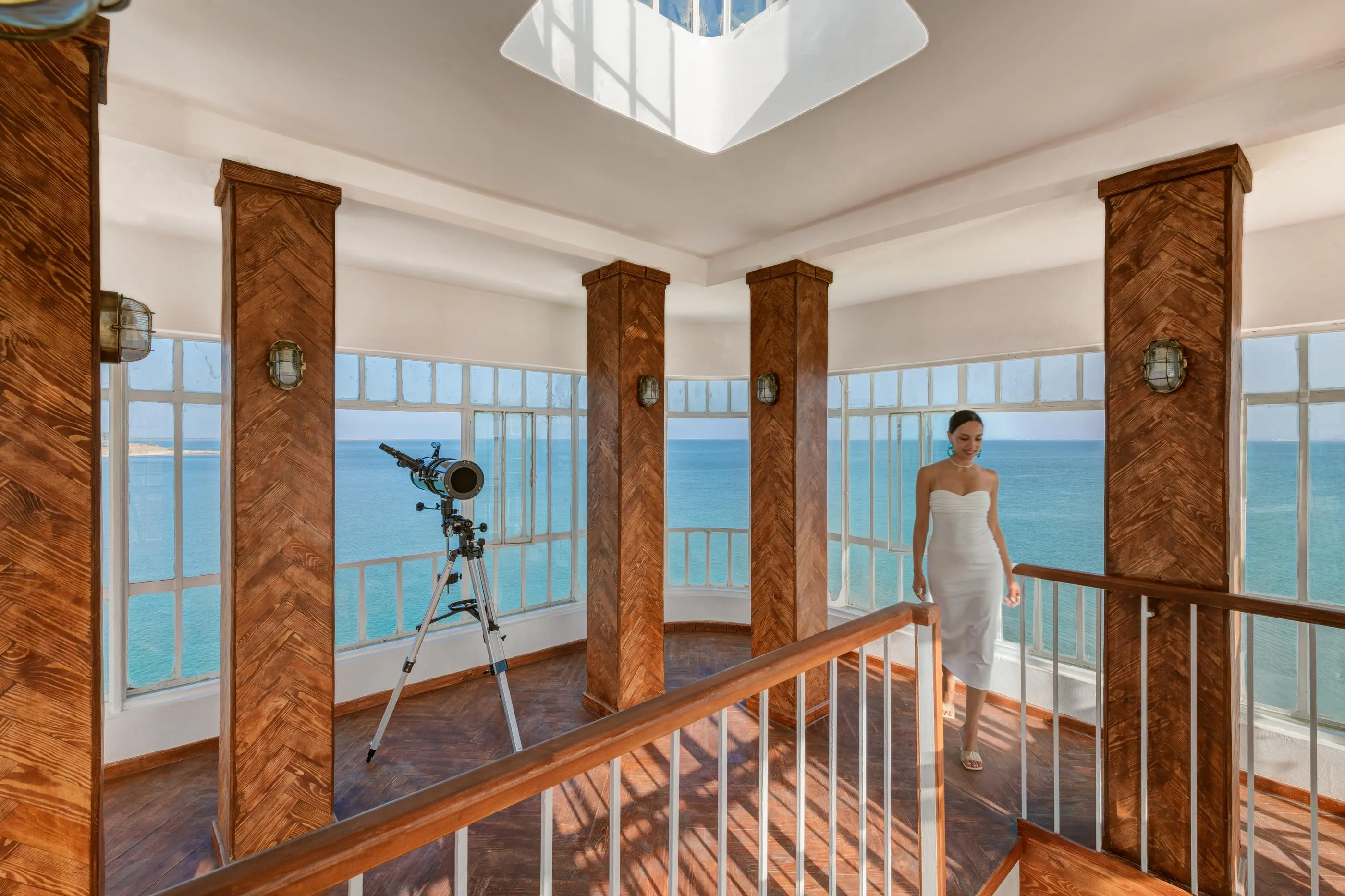 Une femme en robe blanche marche sur un balcon en bois avec vue sur la mer, un télescope est installé près de la rambarde