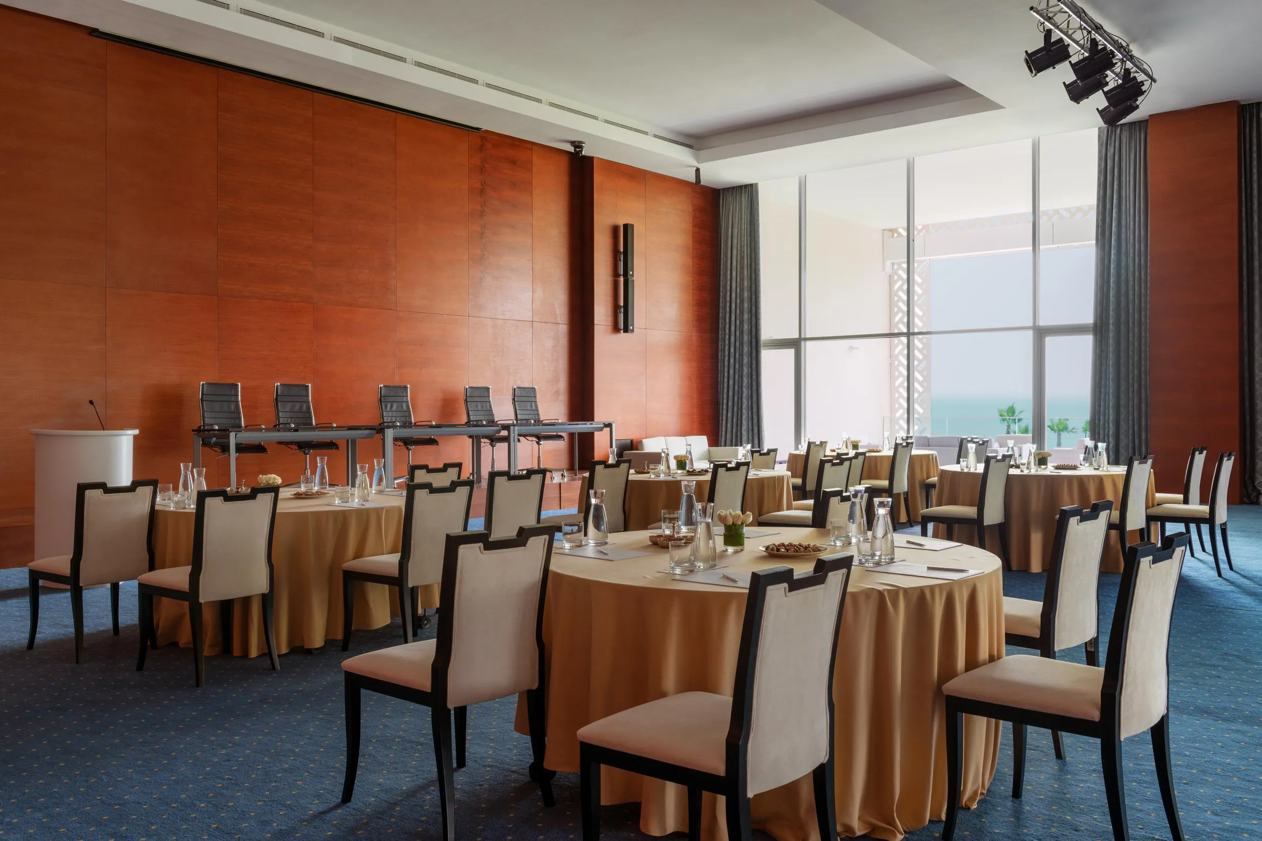 Salle de réunion avec plusieurs tables rondes recouvertes de nappes beiges, des chaises autour et une grande fenêtre avec des rideaux