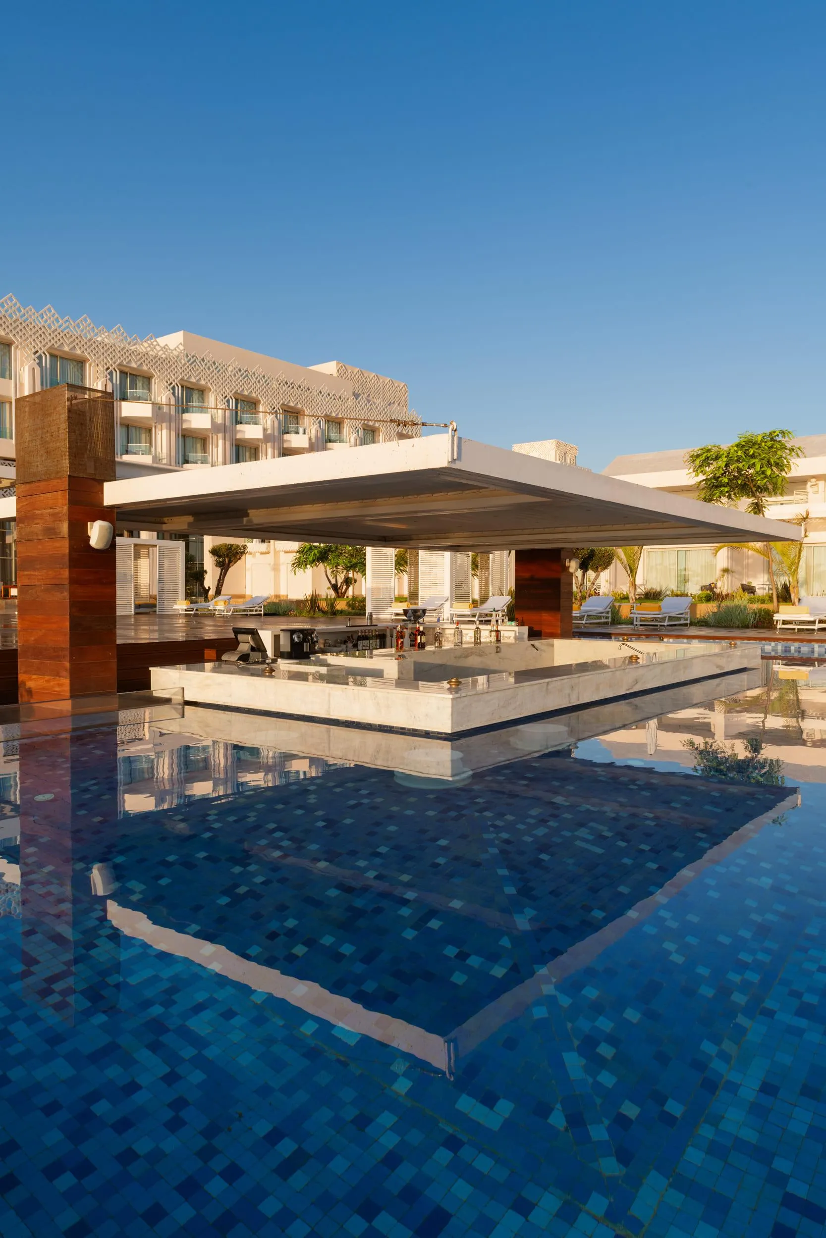 Piscine avec un bar central et des transats autour, reflétant les bâtiments et le ciel bleu clair