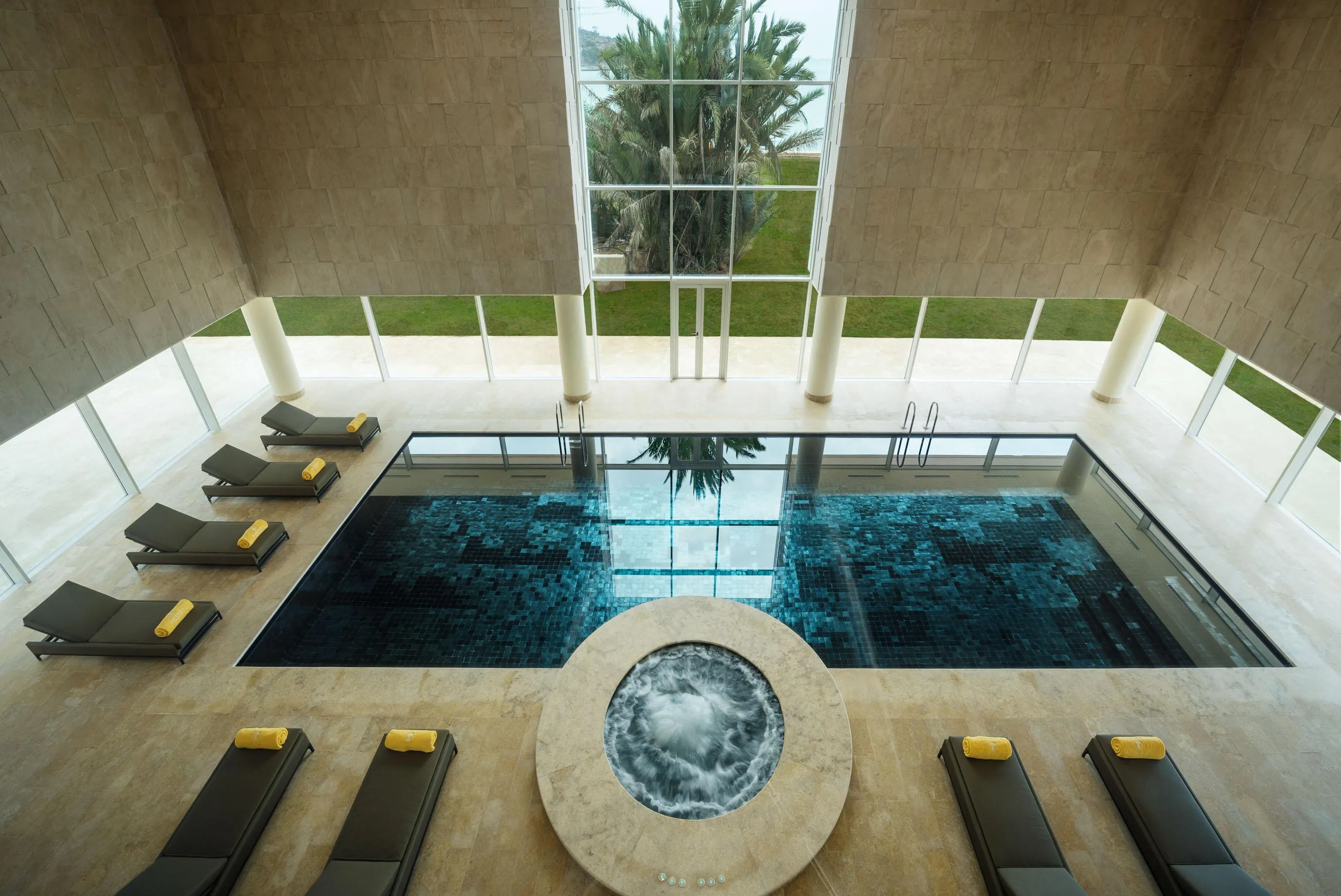 Piscine intérieure avec des chaises longues autour, vue depuis le plafond avec une grande fenêtre et des colonnes